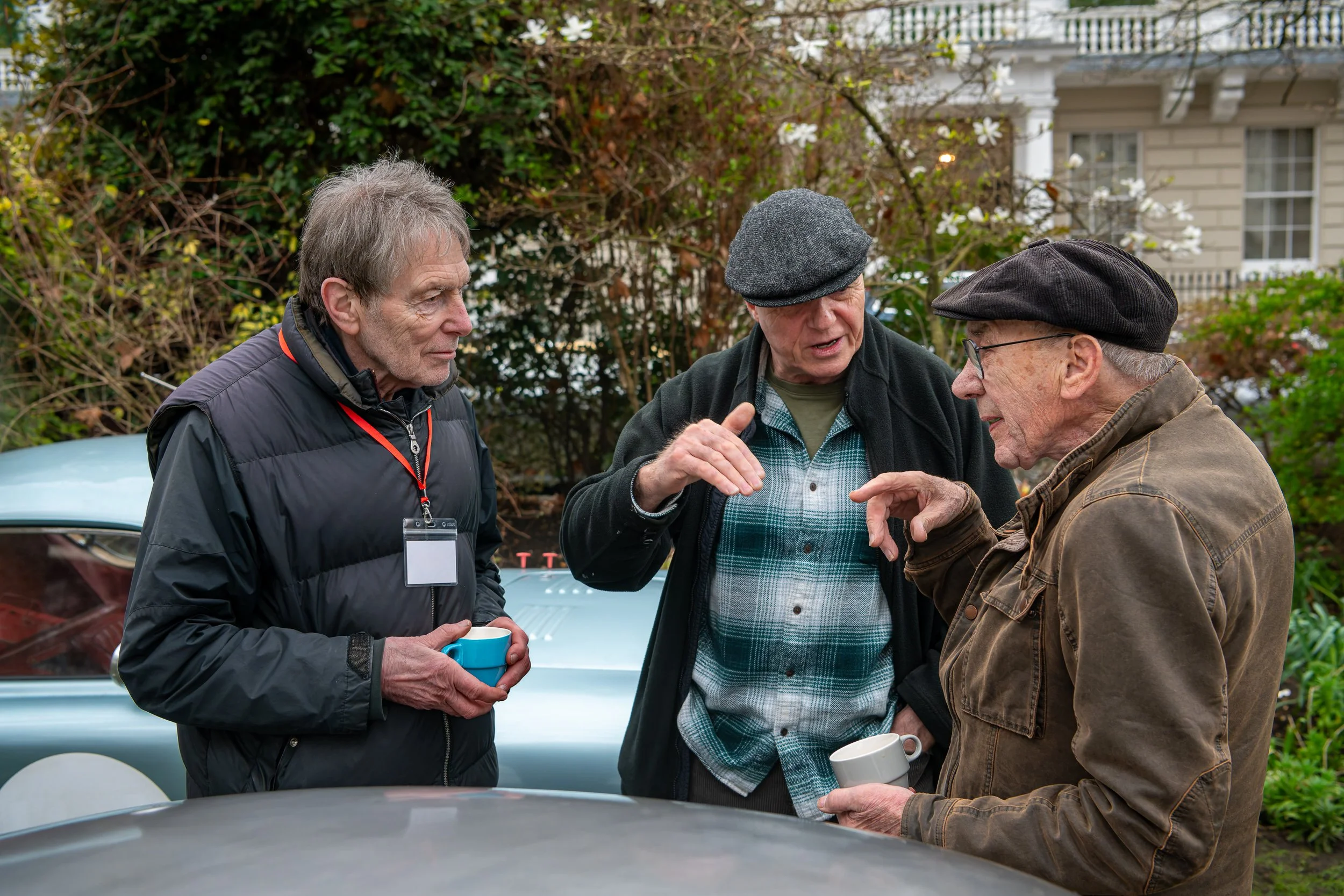 Three elderly men in coats and hats are having a conversation outdoors, holding mugs, with a vintage car and greenery in the background.