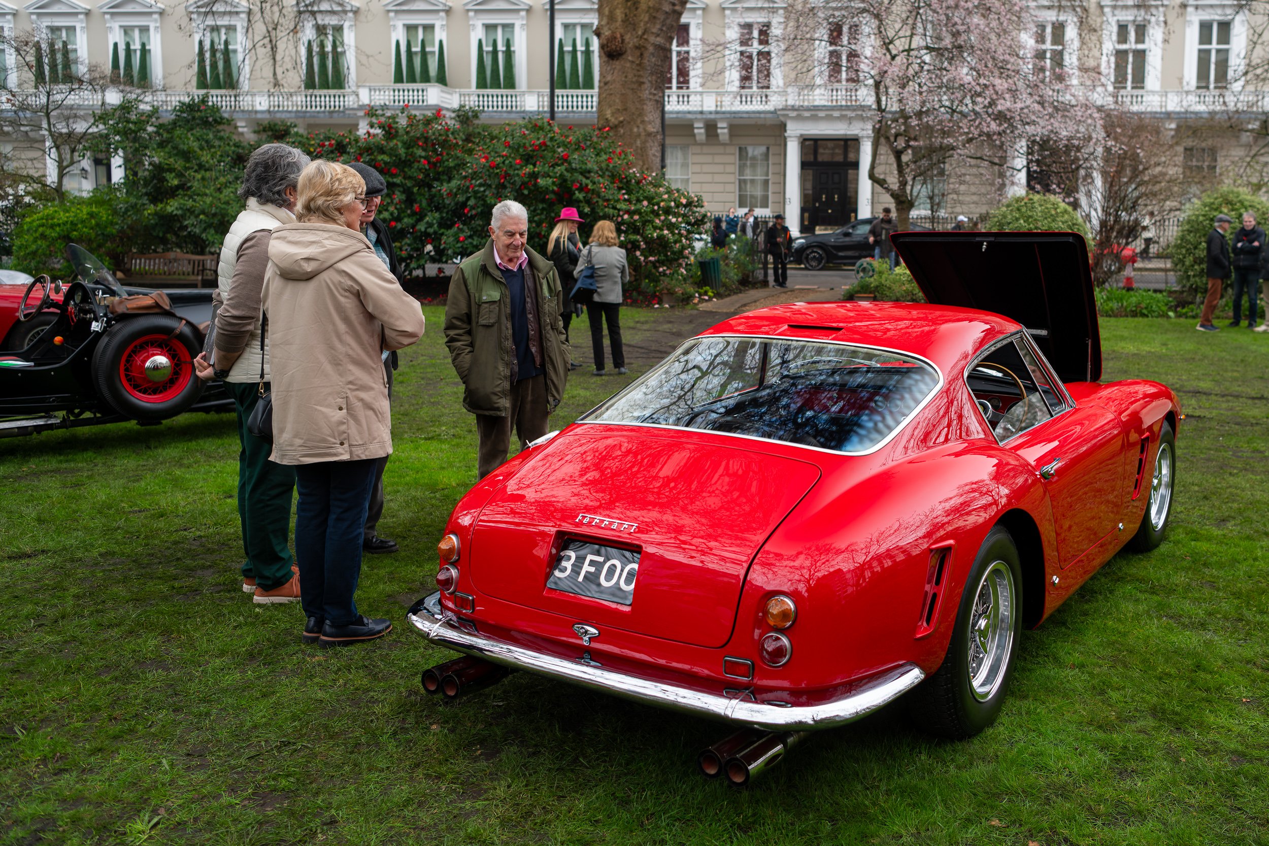 A classic red Ferrari sports car displayed on a grassy area at an outdoor car show, with four people inspecting it closely. The car's rear hatch is open, and others are visible in the background near a grand building.