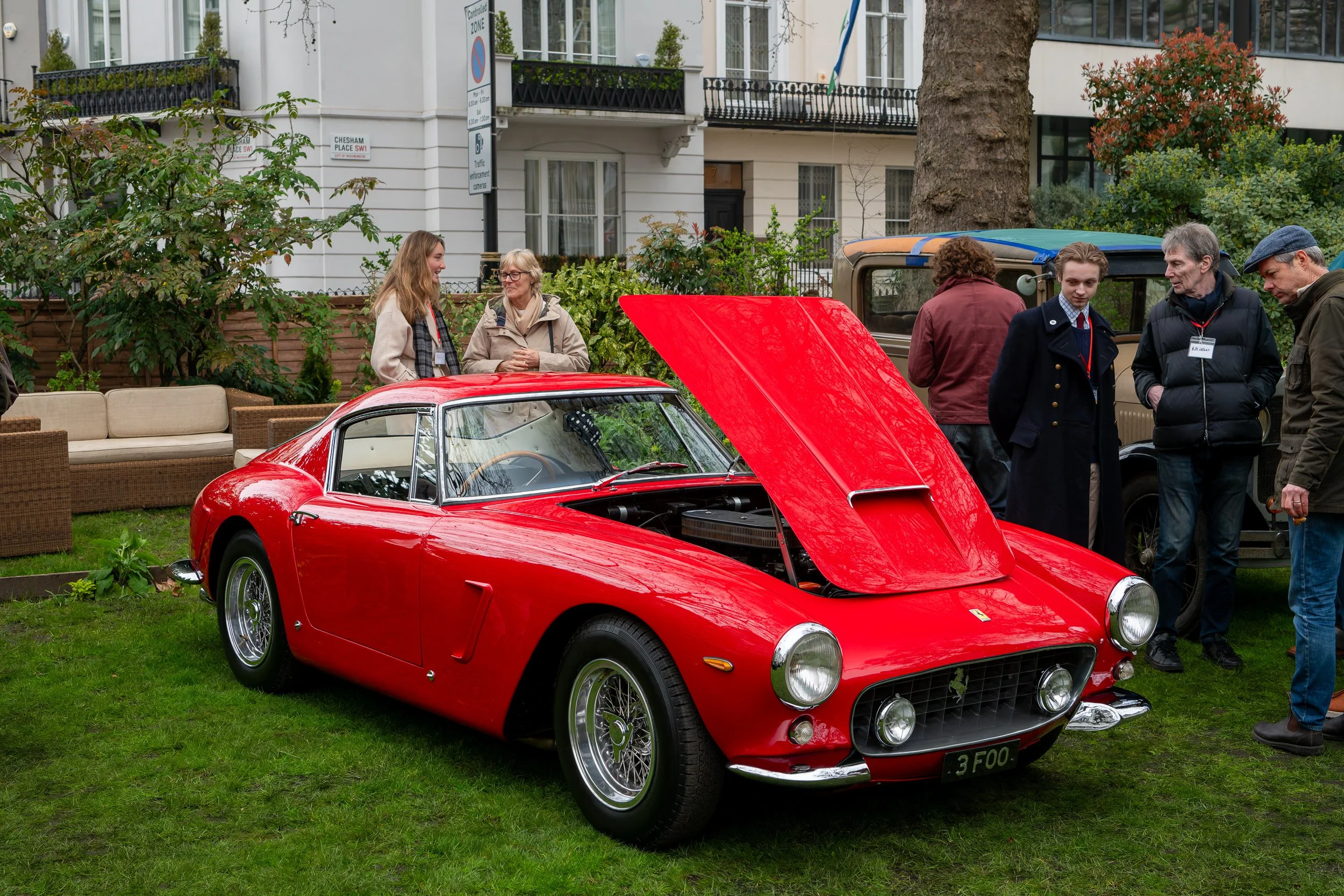 Red vintage Ferrari sports car with its hood open displayed on a grassy area at a car show, surrounded by people looking at the car and talking.
