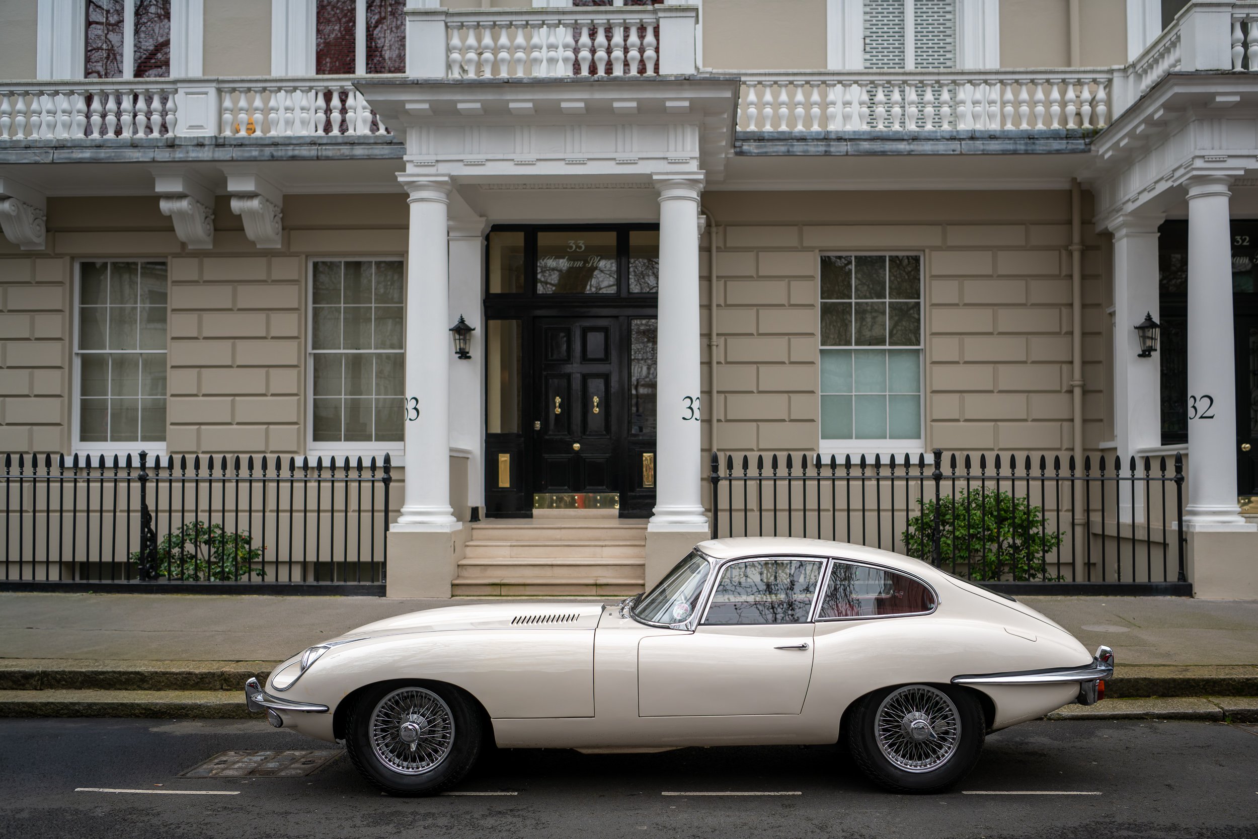 A vintage white car parked in front of a beige residential building with white columns and black door, with a black metal fence along the sidewalk.
