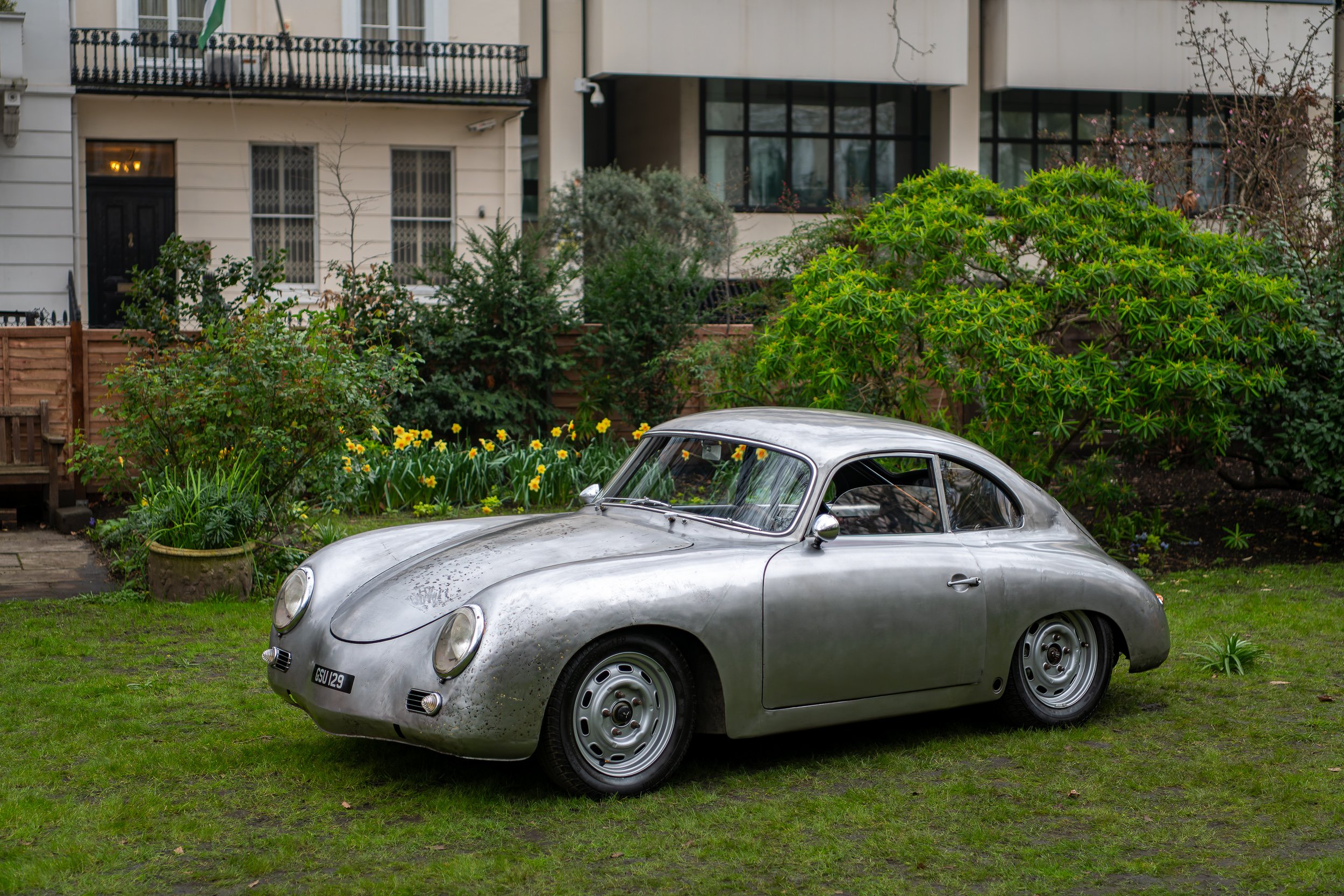 A vintage silver Porsche 356 parked on a grassy lawn in front of a garden with green plants, yellow flowers, and trees, with residential buildings in the background.