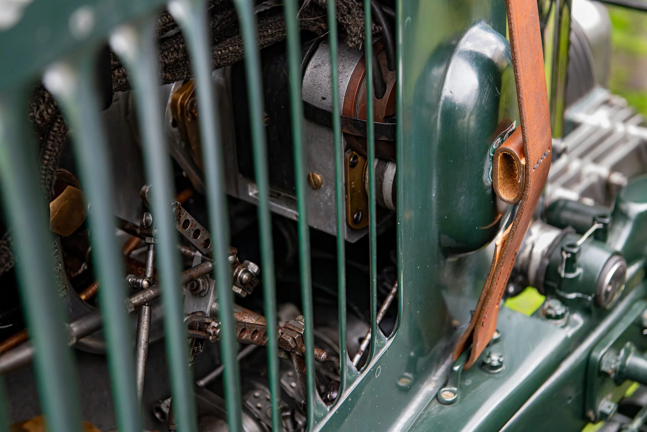 Close-up of an old military or vintage vehicle engine with green body, metal components, and a brown leather strap securing part of the engine, viewed through a green metal grille.