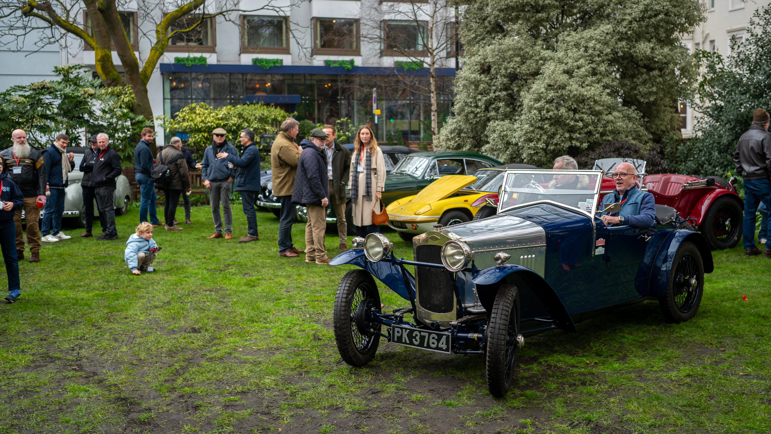 People gathered around vintage and classic cars parked on a grassy area, including a blue vintage car in the foreground. A small child squats on the grass, holding a toy, while adults chat and admire the cars. There are trees and a modern building in