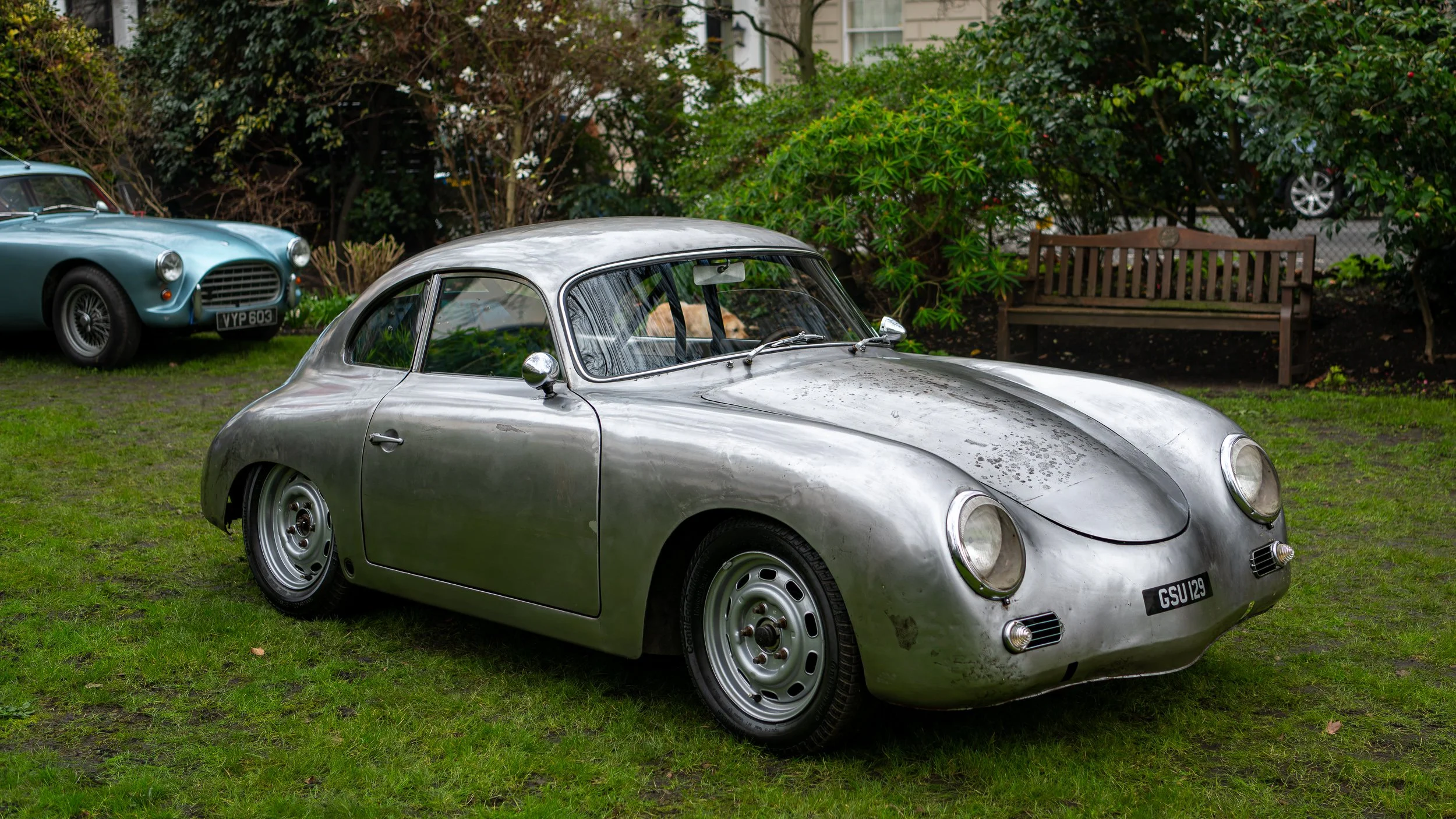 A vintage silver car with dust and dirt on the surface, parked on a grassy area with trees and bushes in the background, alongside another classic blue car.