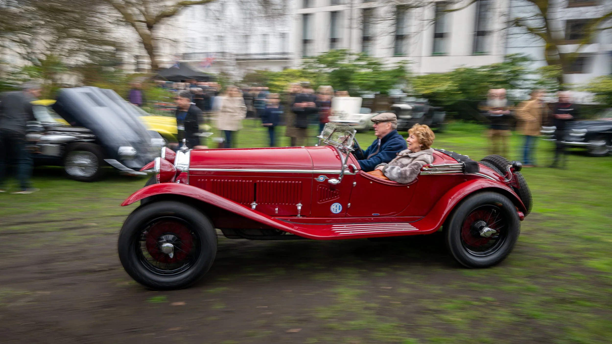 A vintage red convertible car driving through a park with people standing and viewing in the background, some blurred due to motion.