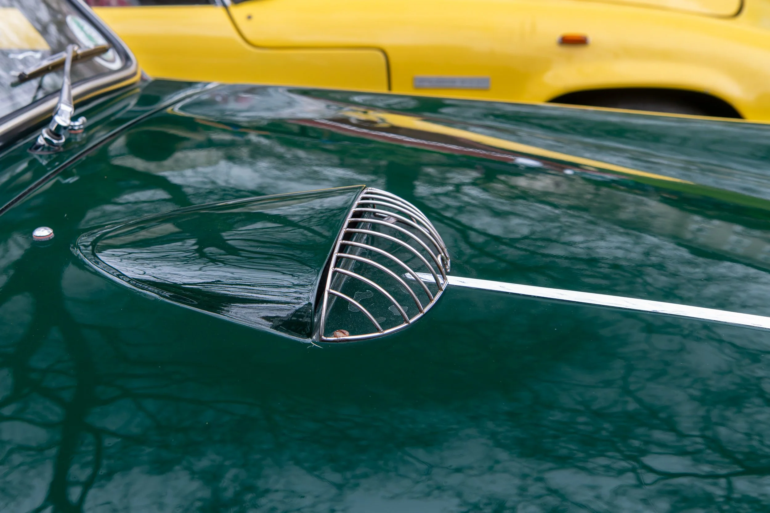 Close-up of a vintage green car with a chrome detail on the hood and a yellow vehicle in the background.