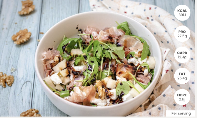 Salad with arugula, sliced pear, walnuts, balsamic glaze, prosciutto, and cheese in a white bowl on a light wooden surface with scattered walnuts and a white napkin with colorful specks.