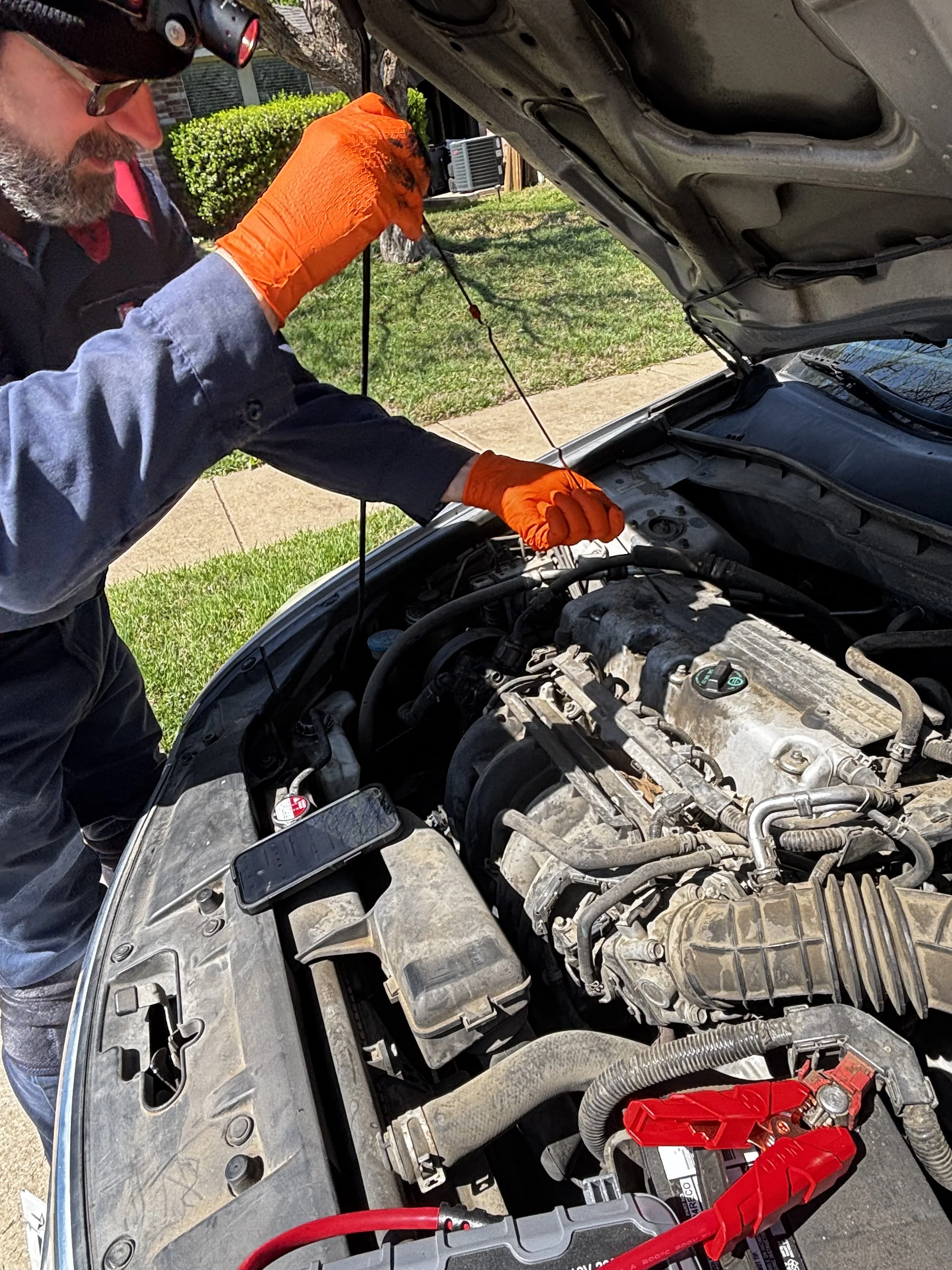 A man wearing orange gloves working on a car engine with the hood open, outdoors on a sunny day.