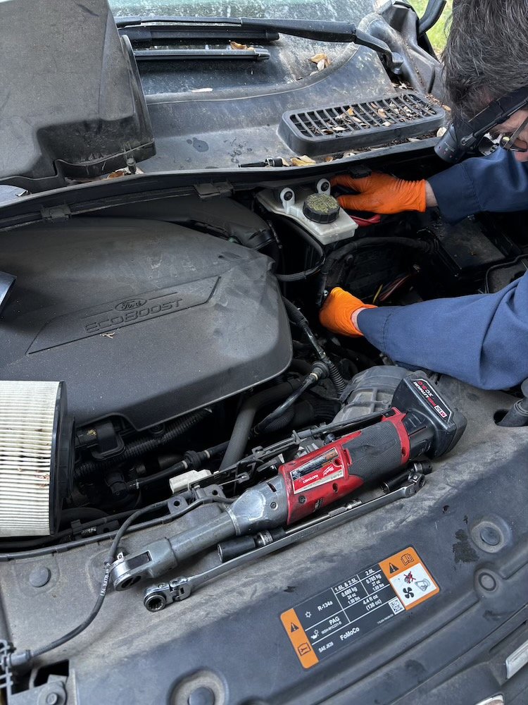 A mechanic wearing orange gloves, glasses, and a blue uniform is using a pneumatic impact wrench in a car's engine bay. The hood is open, revealing engine components and a dirty air filter. The mechanic is working near the windshield area.