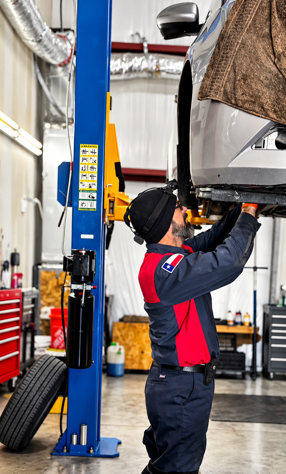 A mechanic wearing glasses, a black beanie, and a Texas flag patch on a navy and red uniform is working underneath a car lifted on a blue hydraulic lift in an auto repair shop. The car appears to be a silver sedan with a brown cloth draped over the front. The shop has various tools, tires, and equipment visible in the background.