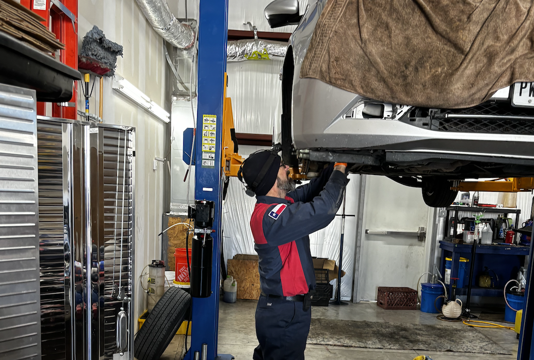 A mechanic wearing a black cap and protective gloves working under a car lifted on a hydraulic lift in a garage. The garage contains various tools, a tire, and storage cabinets.