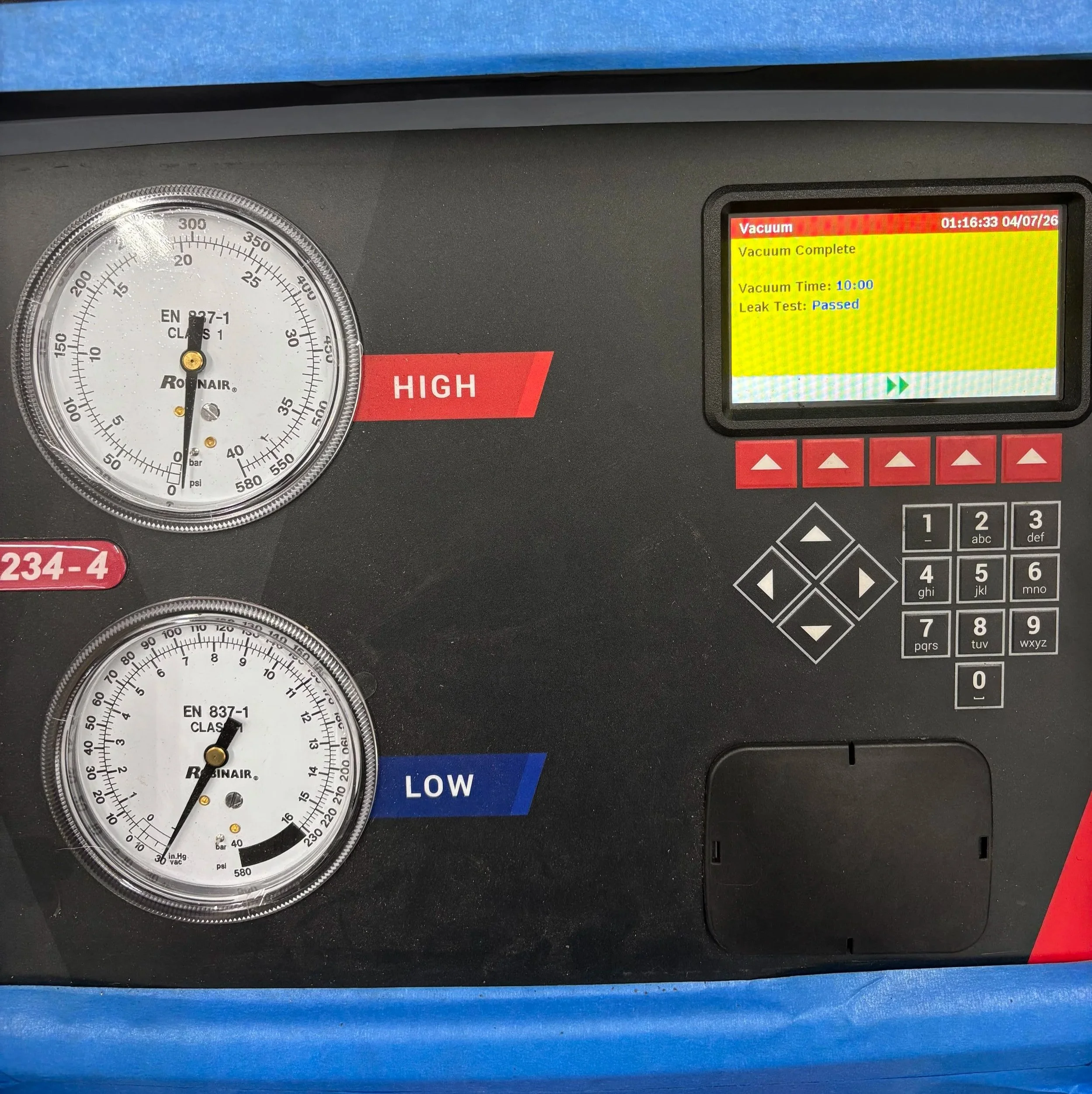 A vehicle air conditioning system service panel displaying gauges and digital screen. The top gauge shows high side pressure at approximately 30 psi. The bottom gauge shows low side pressure at around 10 psi. The digital screen indicates the vacuum process is complete, with a vacuum time of 10 minutes and the leak test status as passed.