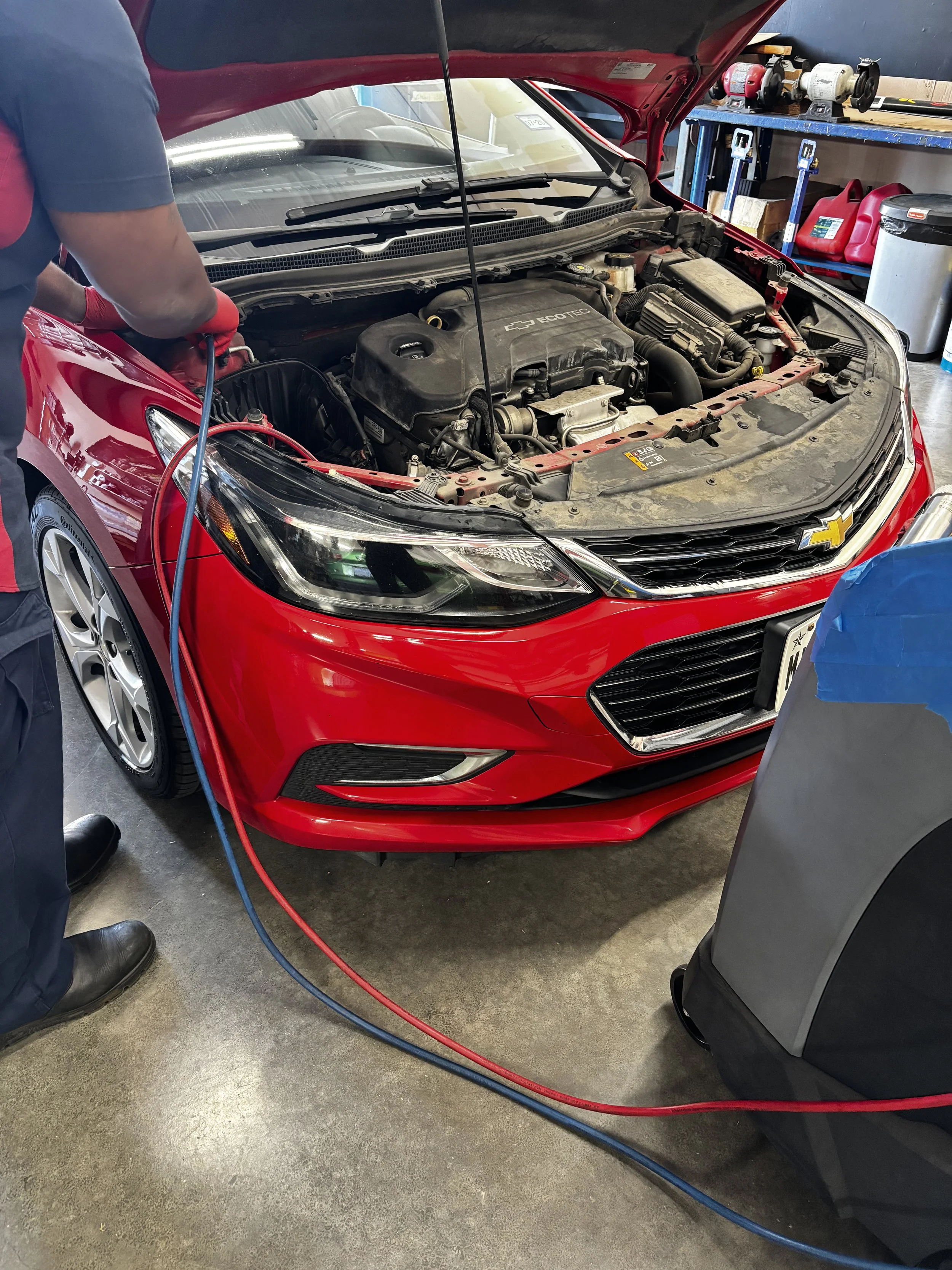 A mechanic providing air conditioning service and inspection on a red Chevrolet car with the hood open, using air conditioning service equipment in an auto repair shop.