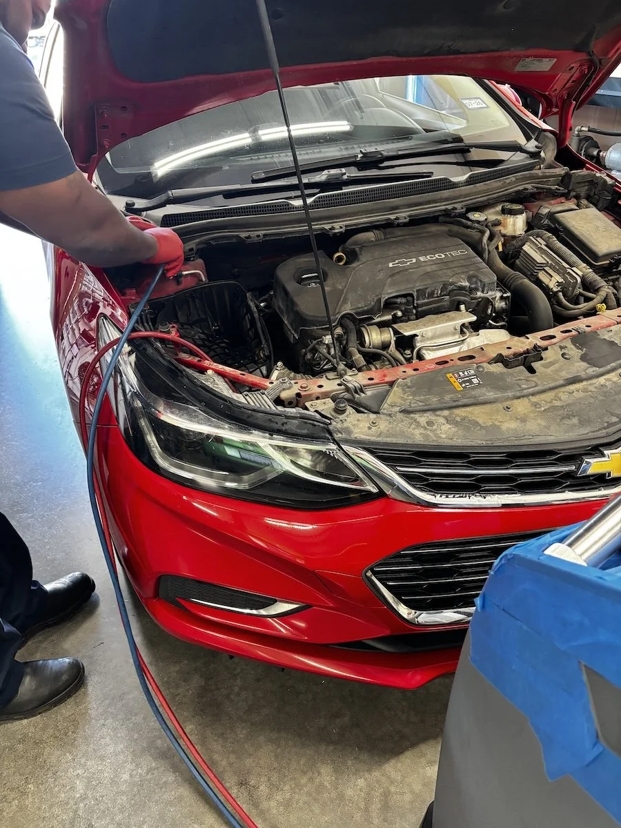 A person working on the engine of a red Chevrolet car with its hood open, using tools while performing maintenance or repairs in a garage.