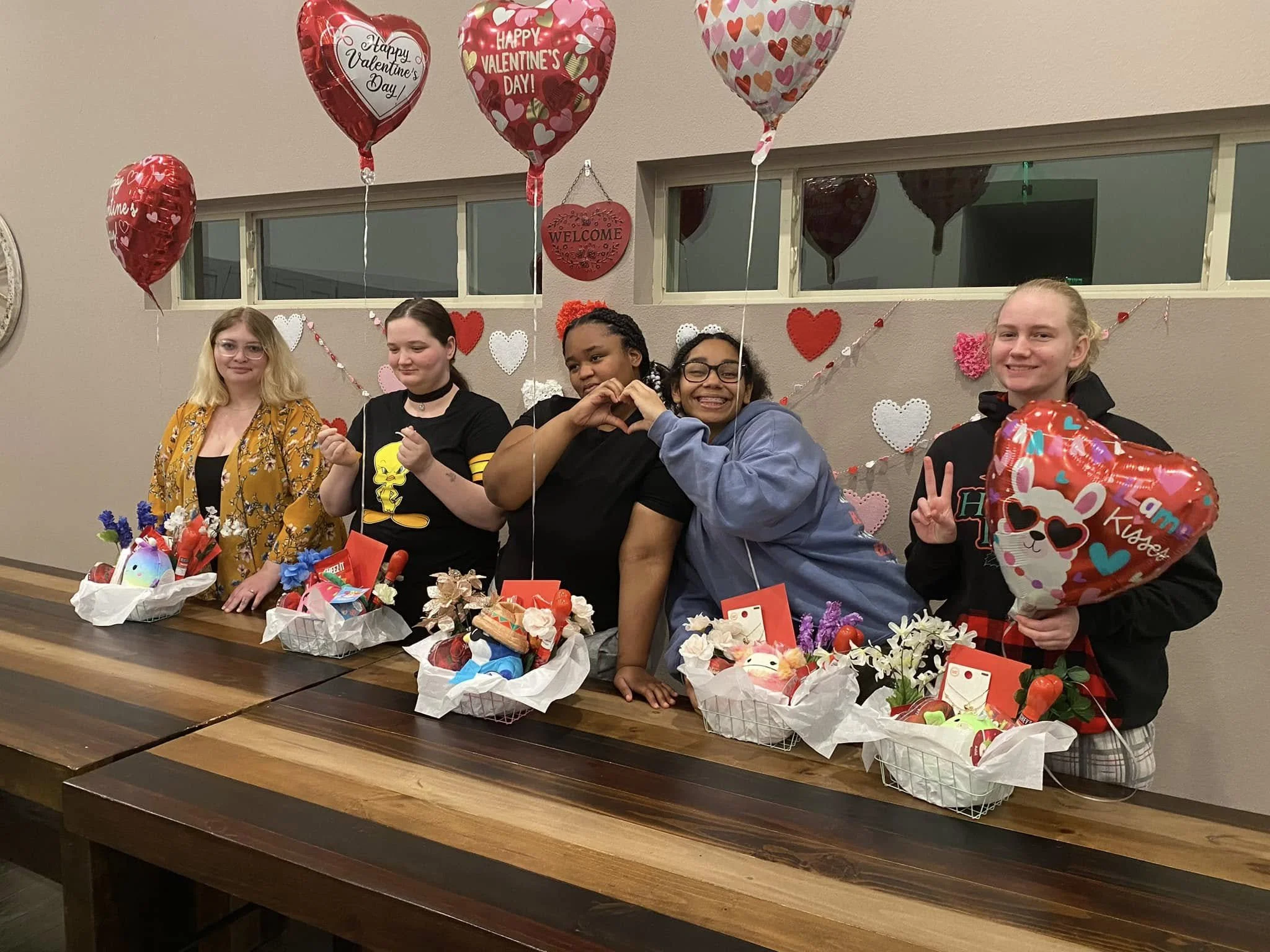 A group of five young women standing behind a long table decorated with Valentine's Day-themed baskets and balloons, with heart-shaped decorations and balloons reading 'Happy Valentine's Day' and 'Welcome' in the background.