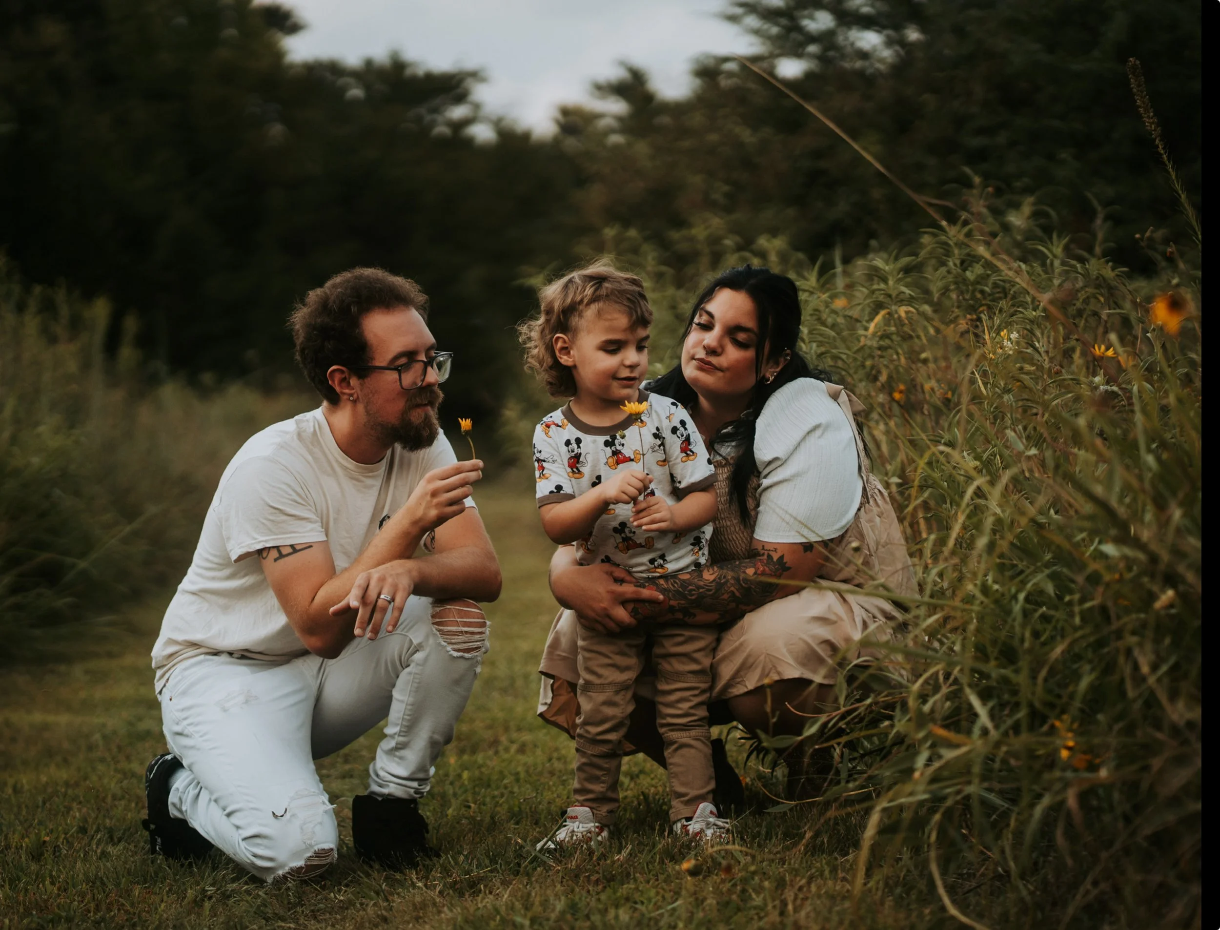 Wichita family of three enjoying a playful outdoor photo session among flowers, with young son interacting with blooms, captured in moody, candid, and affordable photography