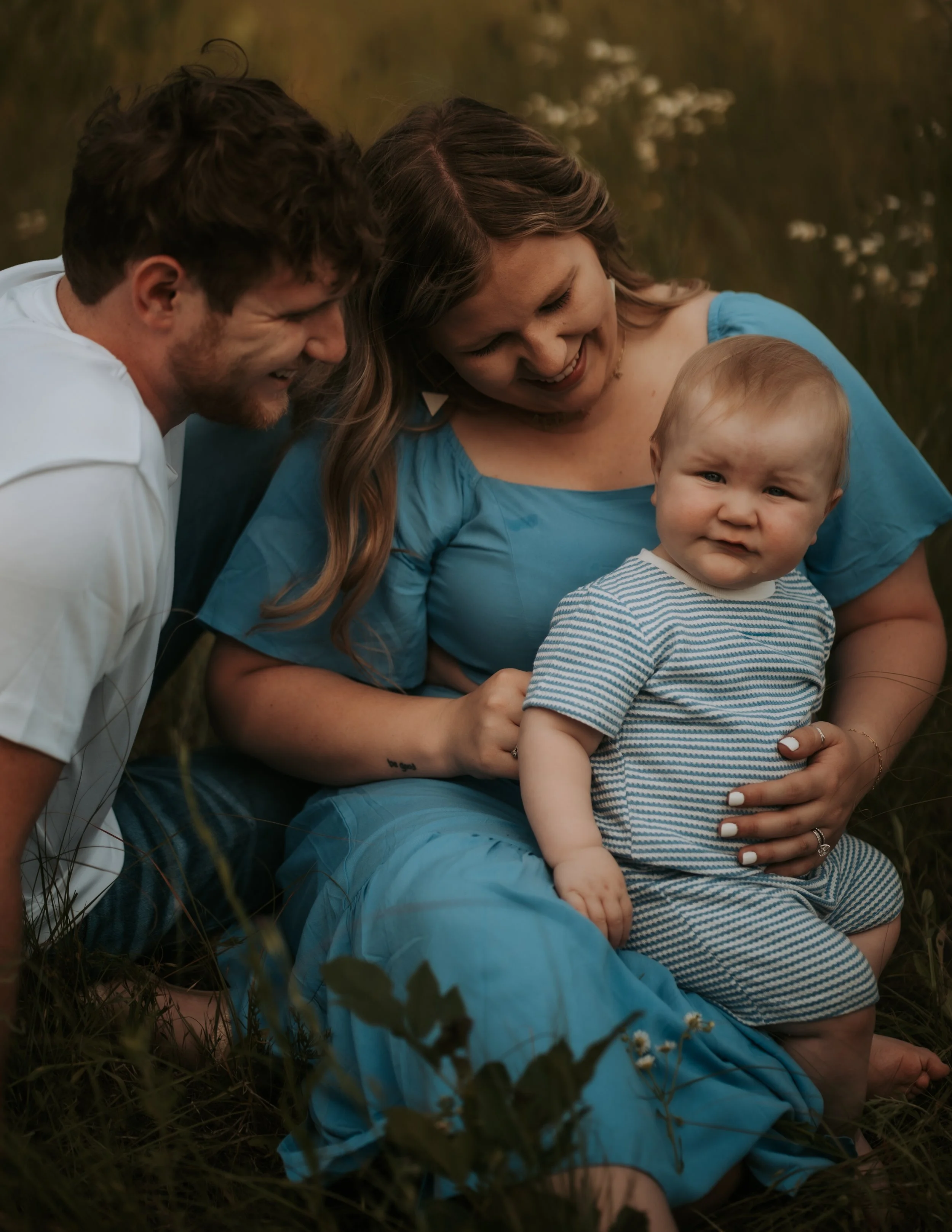 Wichita family portrait with infant sitting in a field, laughing and smiling during a budget-friendly, candid photography session.