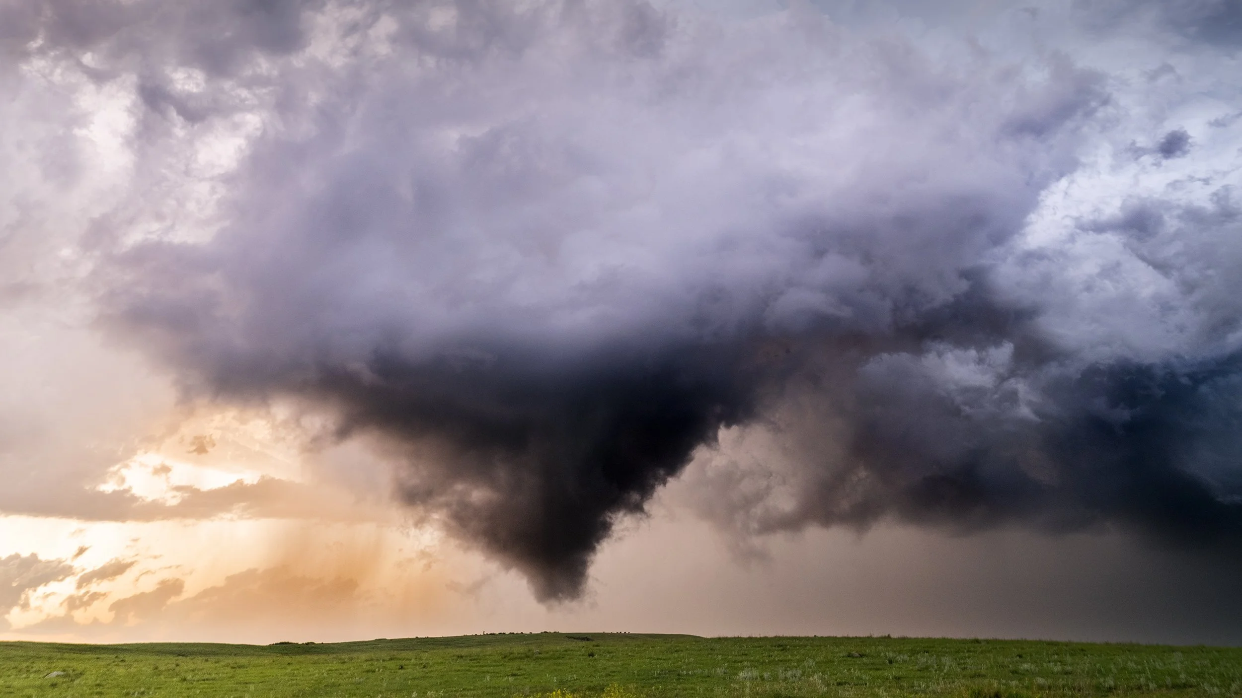 Dark supercell thunderstorm developing over the American Great Plains at sunset, featuring a dramatic rotating wall cloud and near-tornado formation above open prairie grassland. Captured during a storm chasing expedition, this atmospheric severe wea