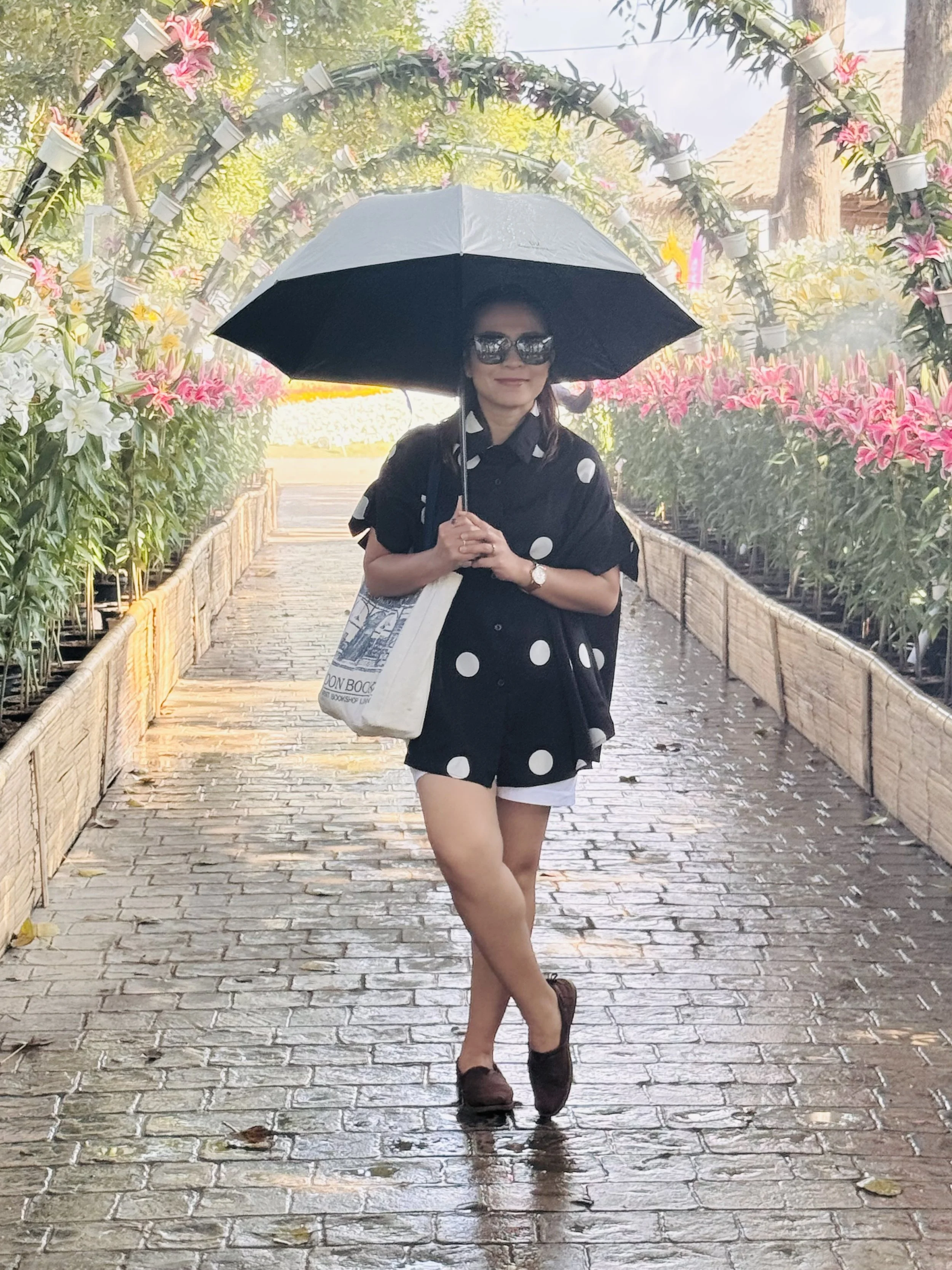 A woman standing on a wet brick pathway under an umbrella, wearing a black polka dot shirt and shorts, sunglasses, with pink lilies and floral archway in the background.