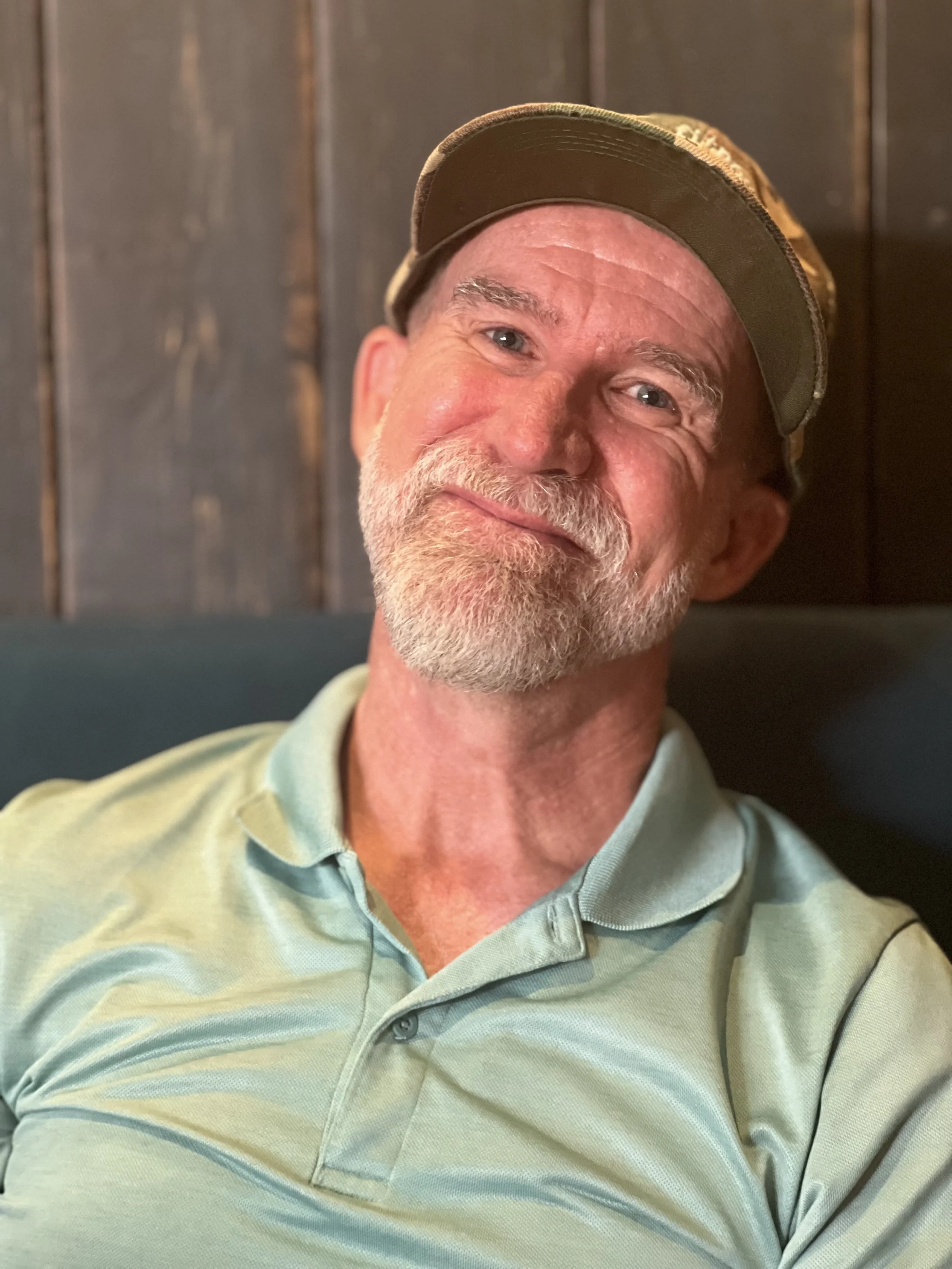 A smiling middle-aged man with a beard and mustache, wearing a cap and a light green polo shirt, sitting against a wooden wall.