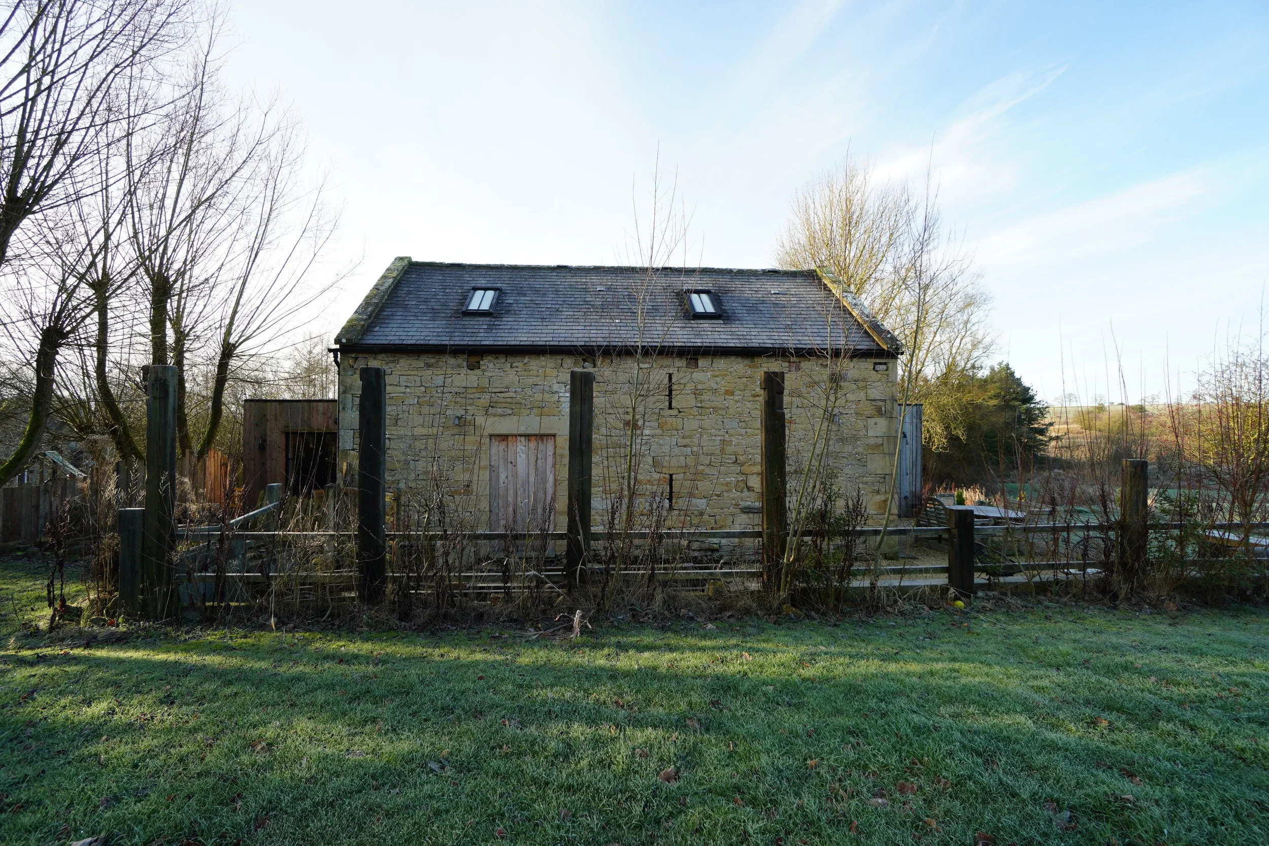A stone house with a sloped roof and two skylight windows, surrounded by a garden with trees and a wooden fence, under a partly cloudy sky.