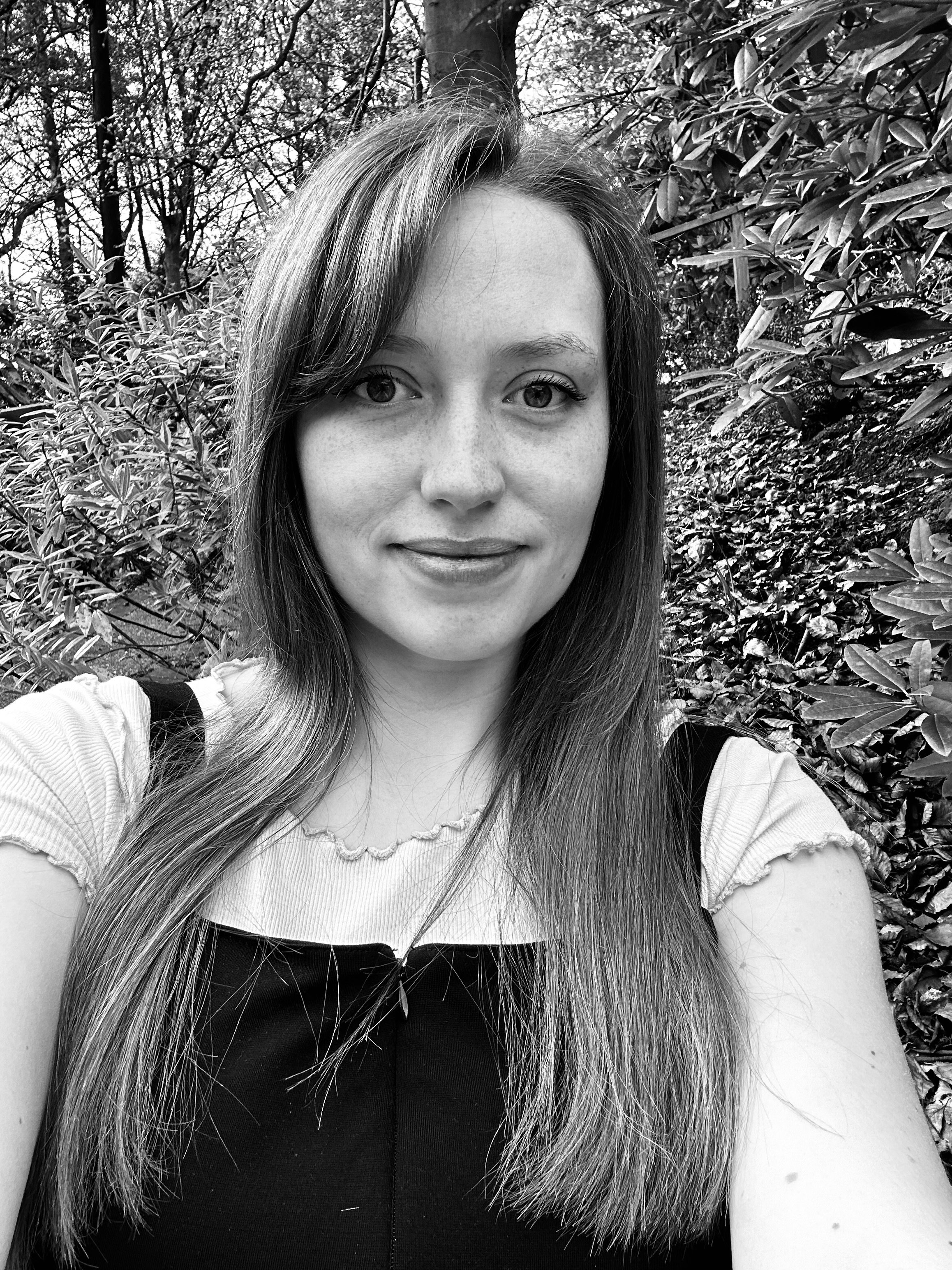 A young woman with long hair smiling outdoors among trees and foliage in black and white.