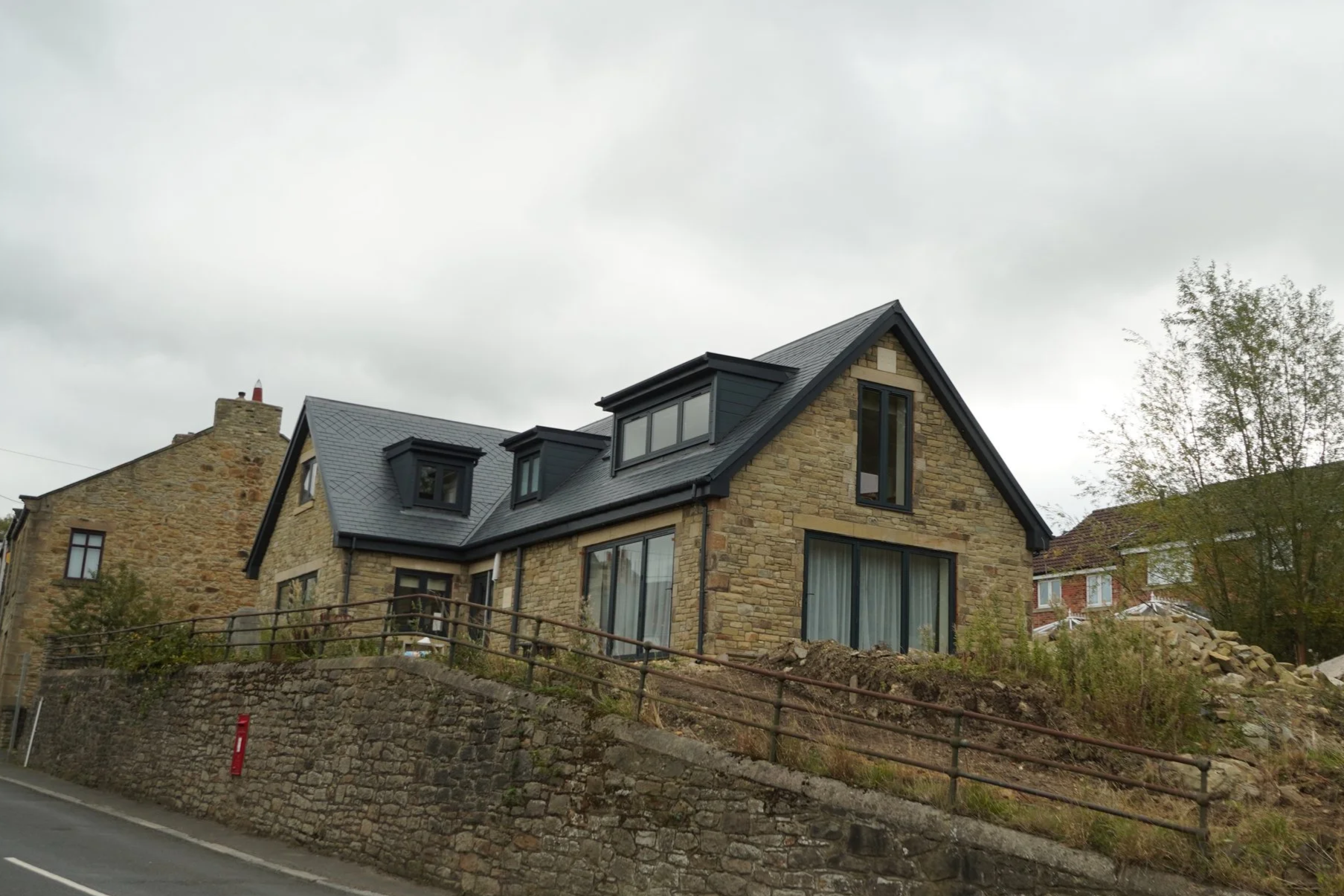 A stone house with black window frames and a black roof, situated on a raised lot with a stone retaining wall and a pathway in front. The house has multiple rooftops, including a gabled roof with dormer windows. Surrounding the house are some trees a