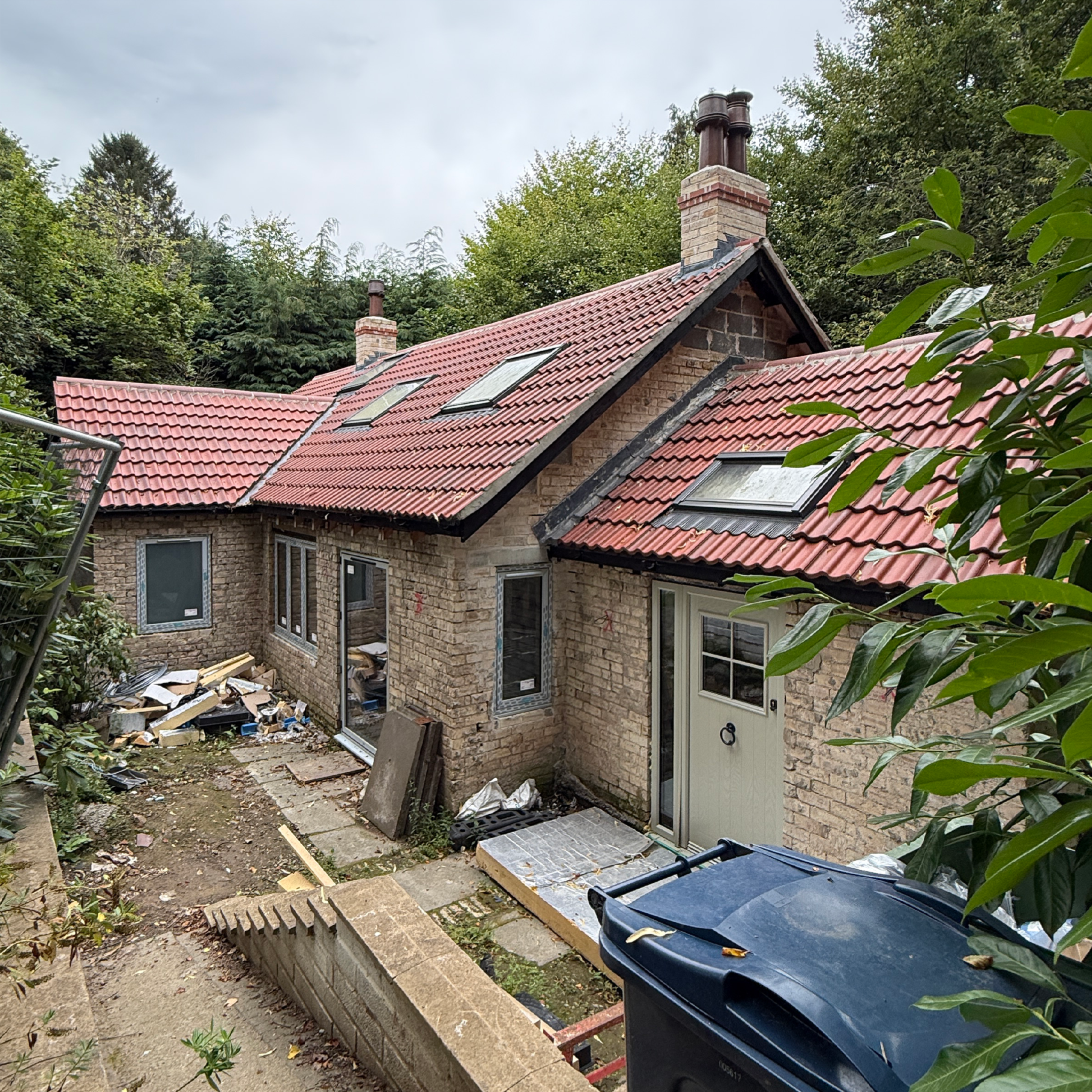 Entrance with brick house, red tile roof, multiple chimneys, and surrounding green trees, with some construction debris and materials in the yard.