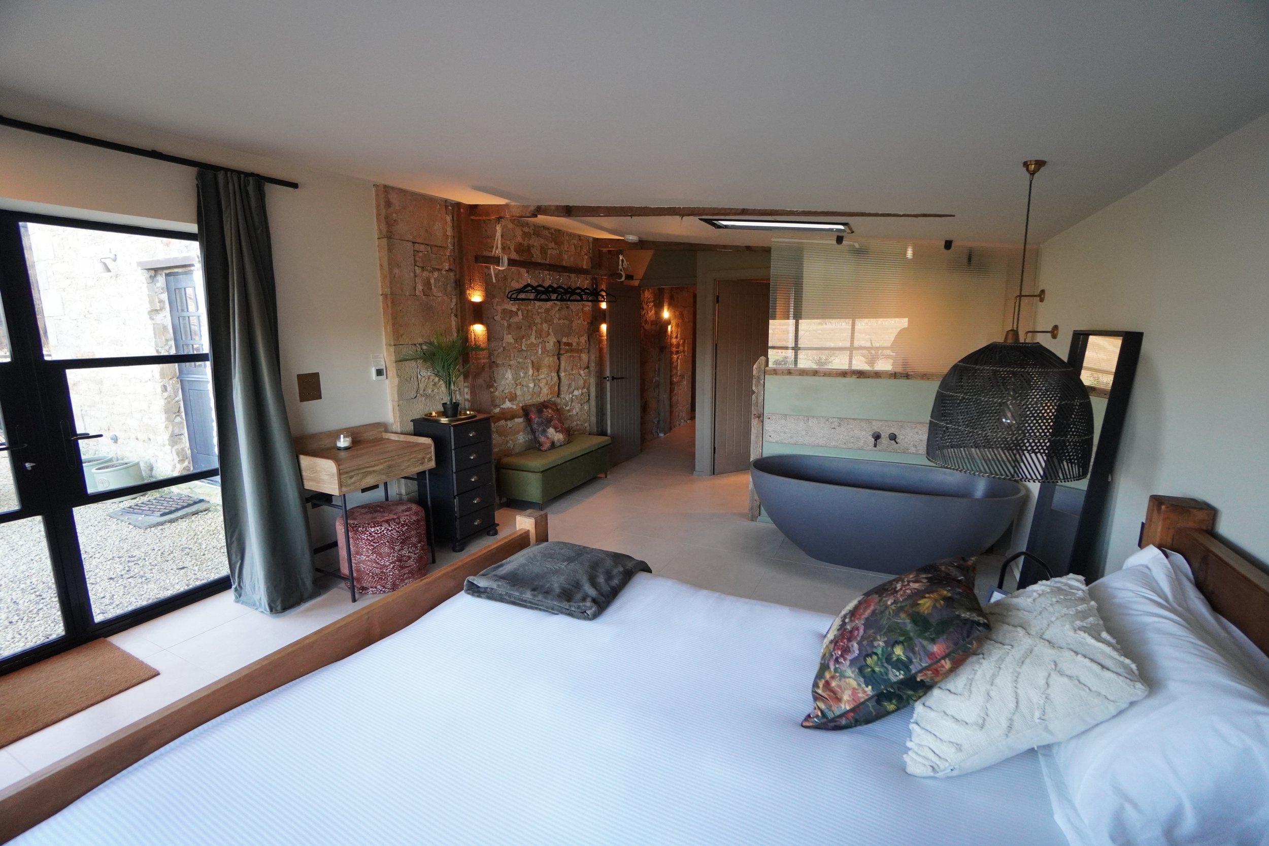 A bedroom with a bed, pillows, and a blanket, next to a window with curtains. There is a small table with a plant and a pillow, a black dresser, and a unique black bathtub. The room has exposed brick wall accents and modern lighting fixtures.