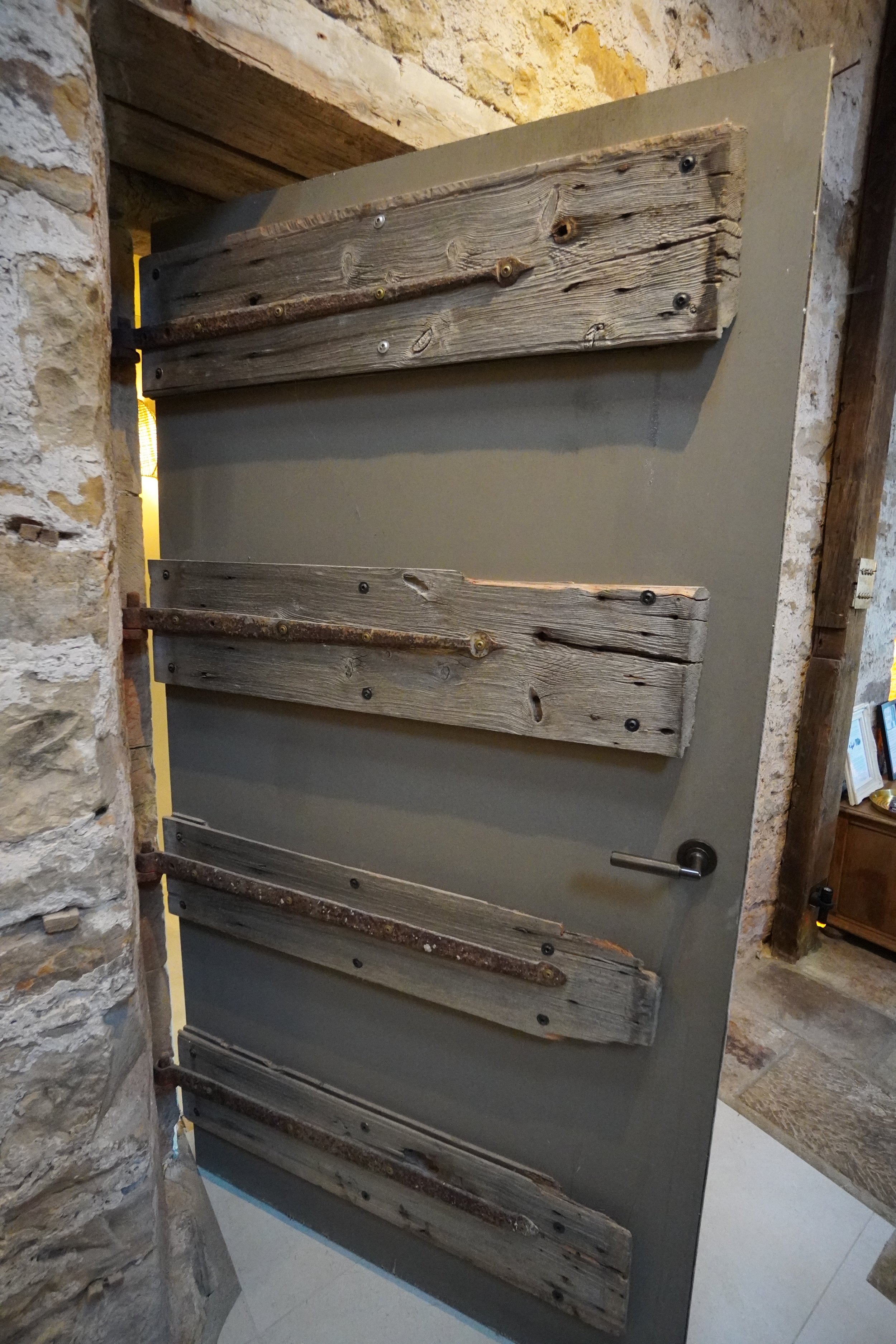 Image of a gray door with wooden planks attached horizontally, equipped with a metallic handle, set in a rustic stone wall.
