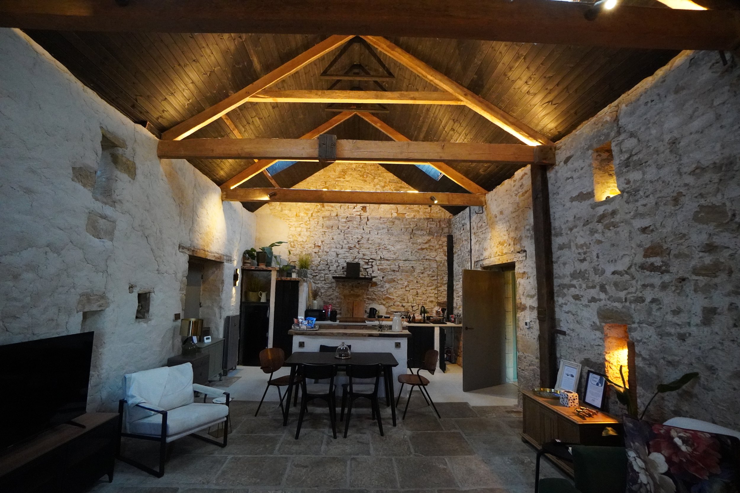 Interior of a rustic living space with exposed stone walls, wooden beams, and a vaulted ceiling. Includes furniture such as chairs, a table, a couch, and decorative plants, with warm lighting.