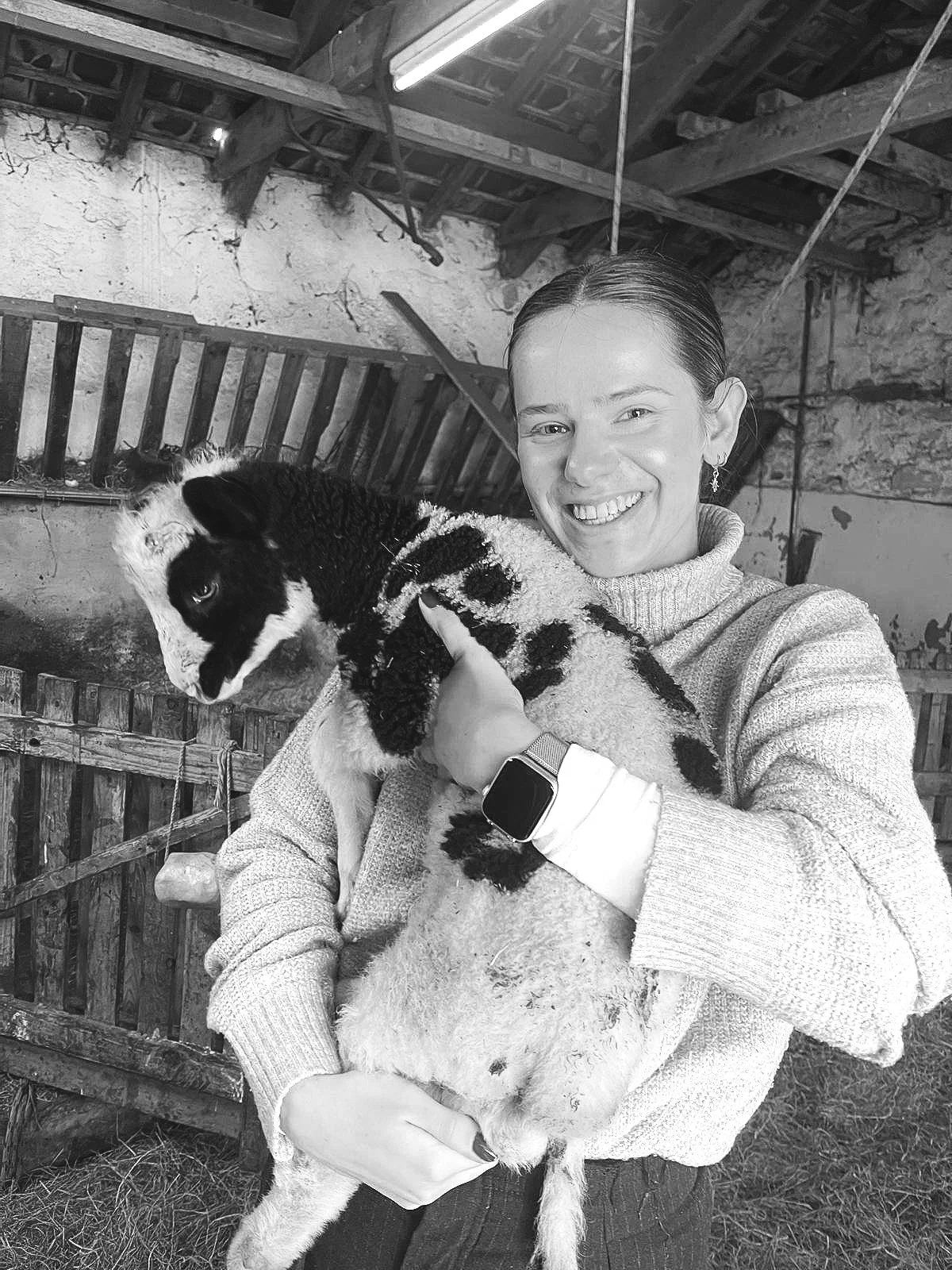 A woman smiling while holding a black and white baby goat inside a rustic barn.