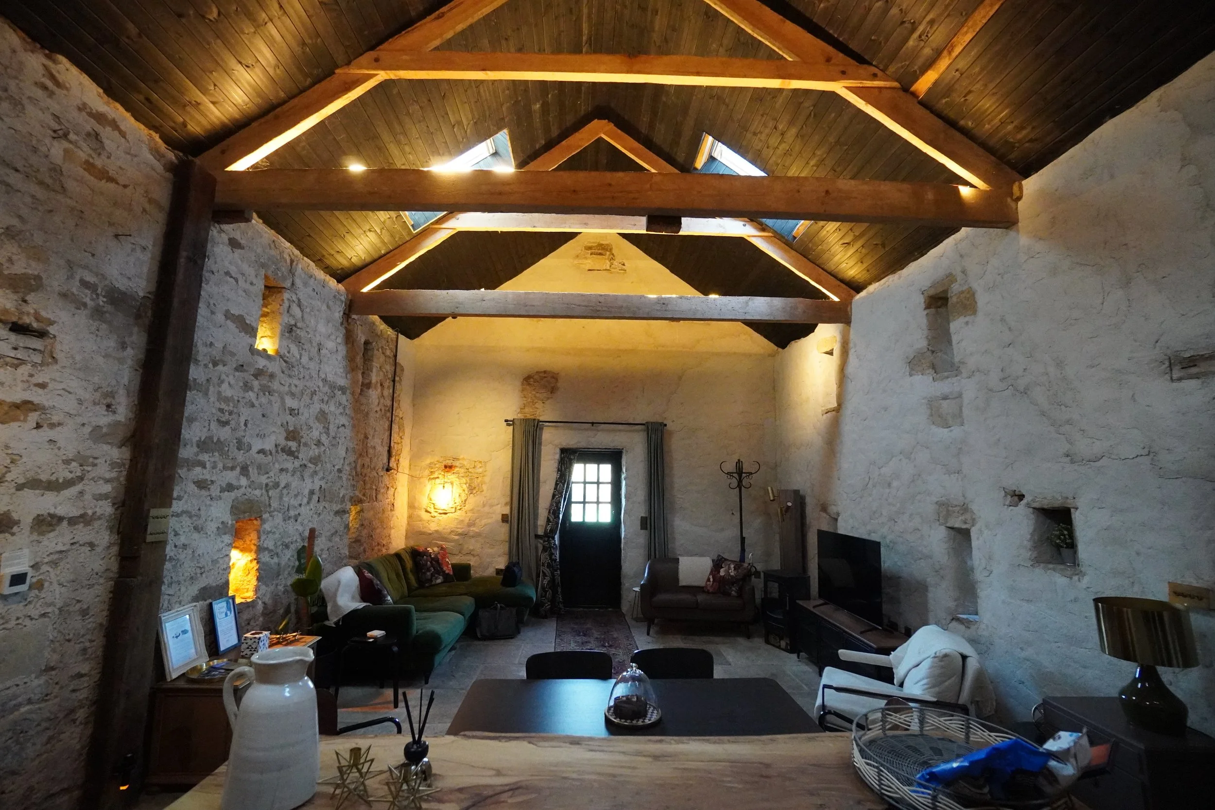 Interior of a rustic living room with stone walls, wooden beams, and a vaulted ceiling. There are sofas, a coffee table with pictures and decorative items, and a window with curtains. Warm lighting illuminates the space.