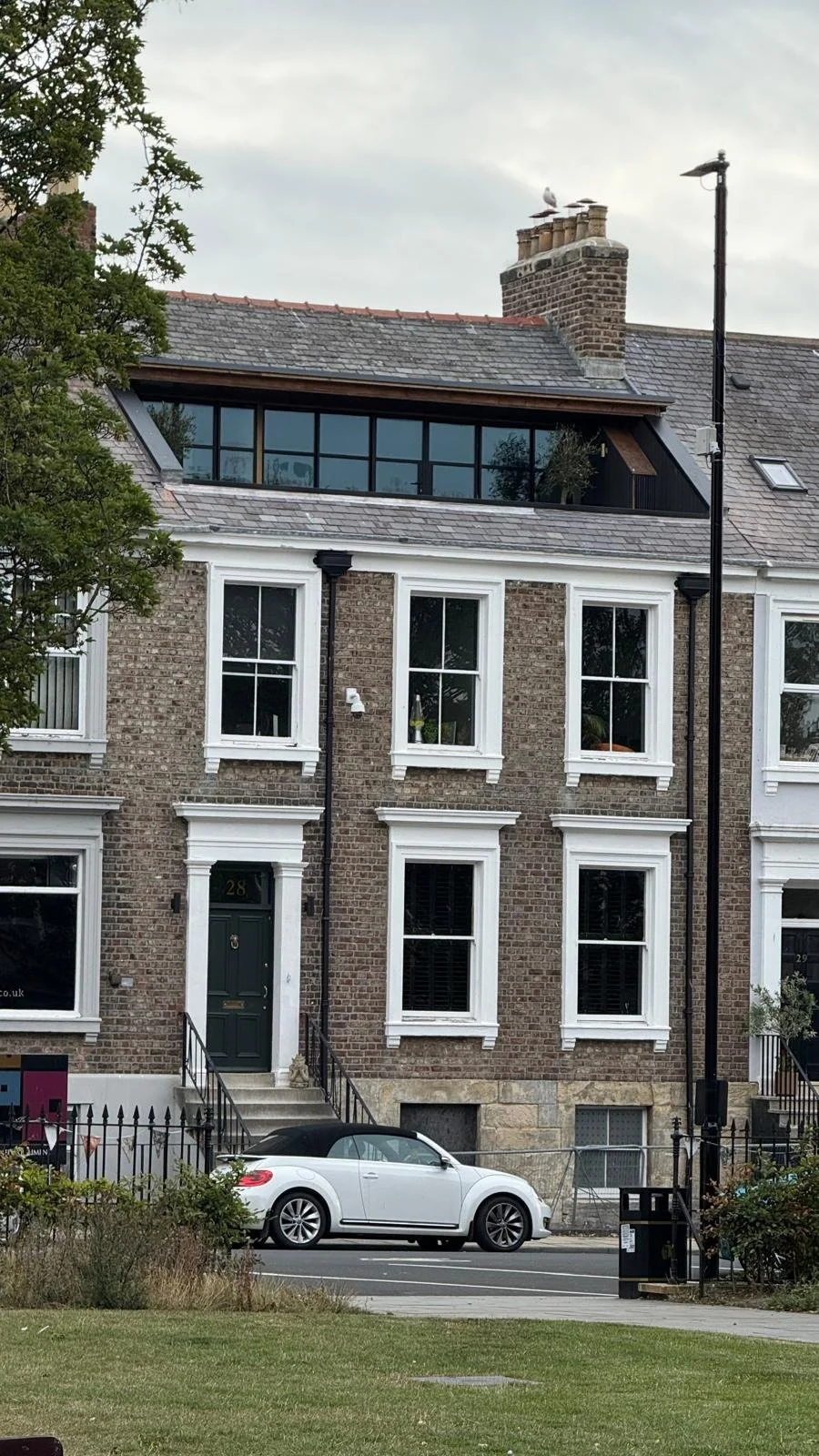 A multi-story brick residential building with white window frames, a green front door, and black railings. A white convertible car is parked in front, and a tree is on the left side. The roof has an attic window and a chimney with a bird perched on t