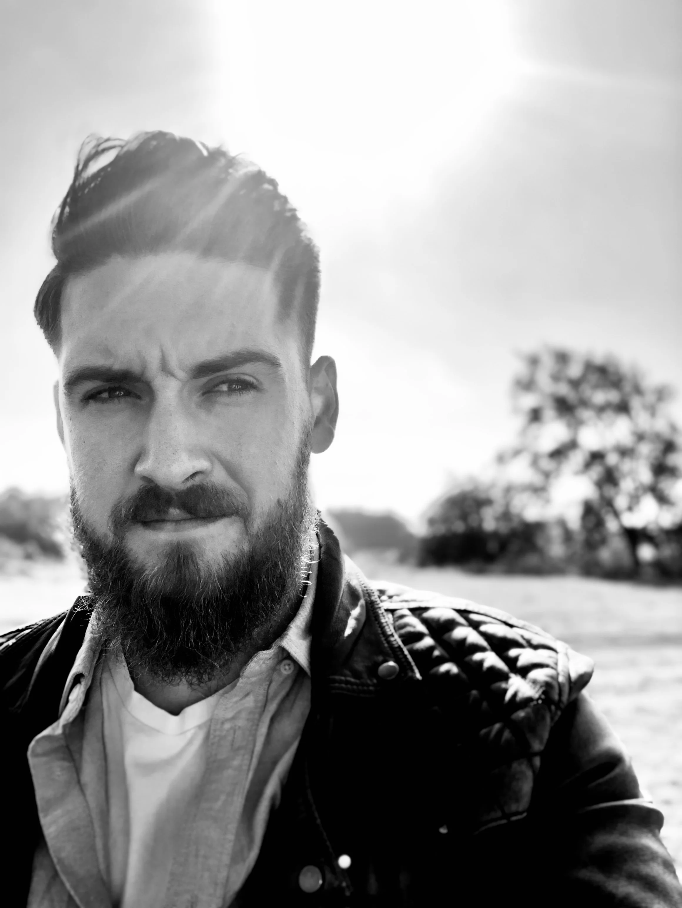 Black and white photo of a bearded man with styled hair, wearing a leather jacket over a collared shirt, outdoors with a tree and open field in the background under bright sunlight.