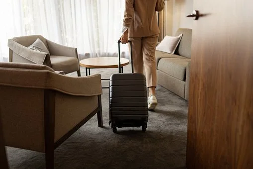 Person with a small rolling suitcase walking through a living room with armchairs and a coffee table.