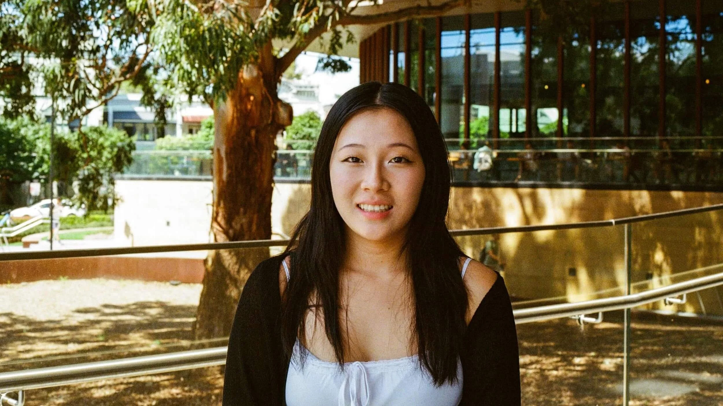 A young woman with long dark hair smiling outdoors on a sunny day, standing near a railing with a tree and modern building in the background.