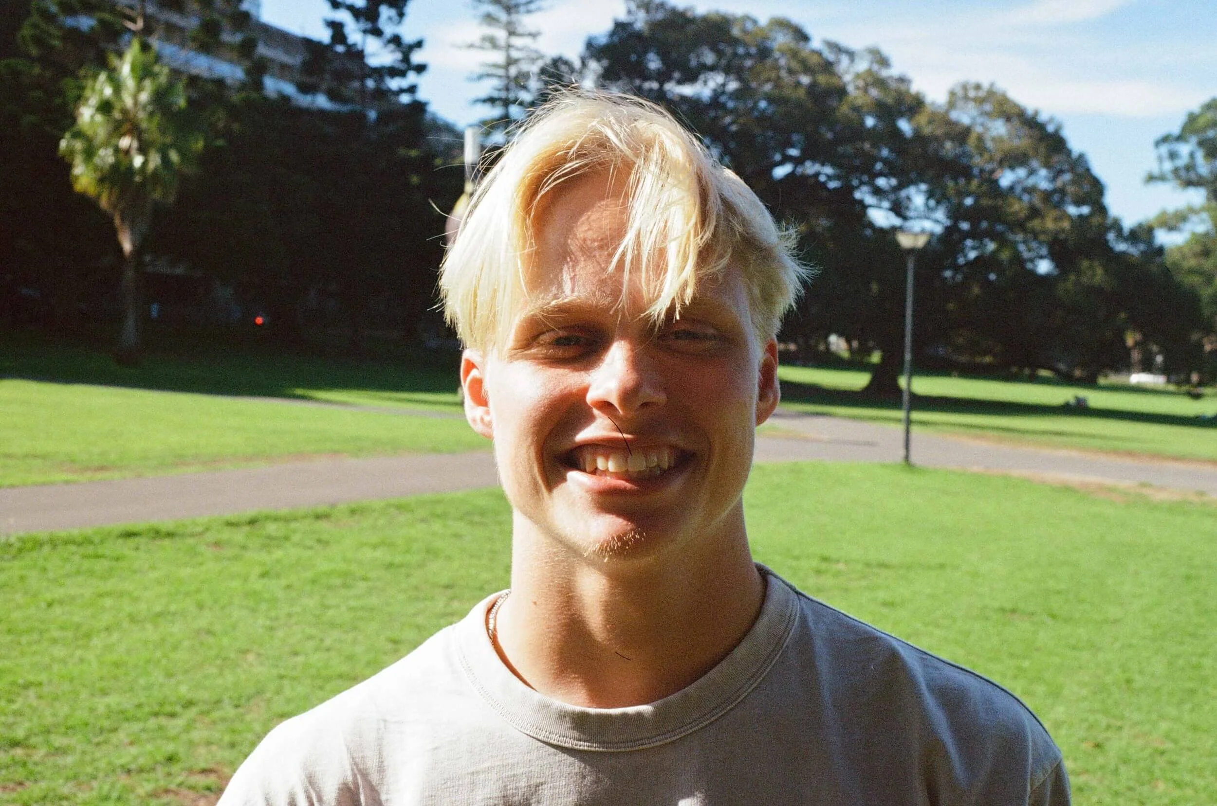 A young man with blonde hair smiling, standing in a park with trees, grass, and a pathway in the background.