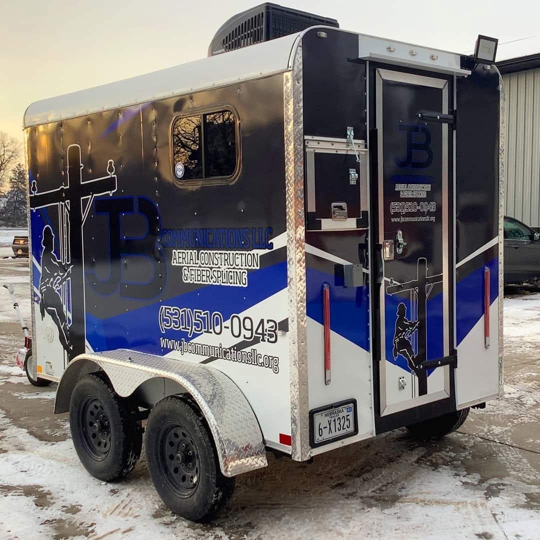 A service trailer for JB Communications LLC, specializing in aerial construction and fiber splicing, parked on snow-covered ground.