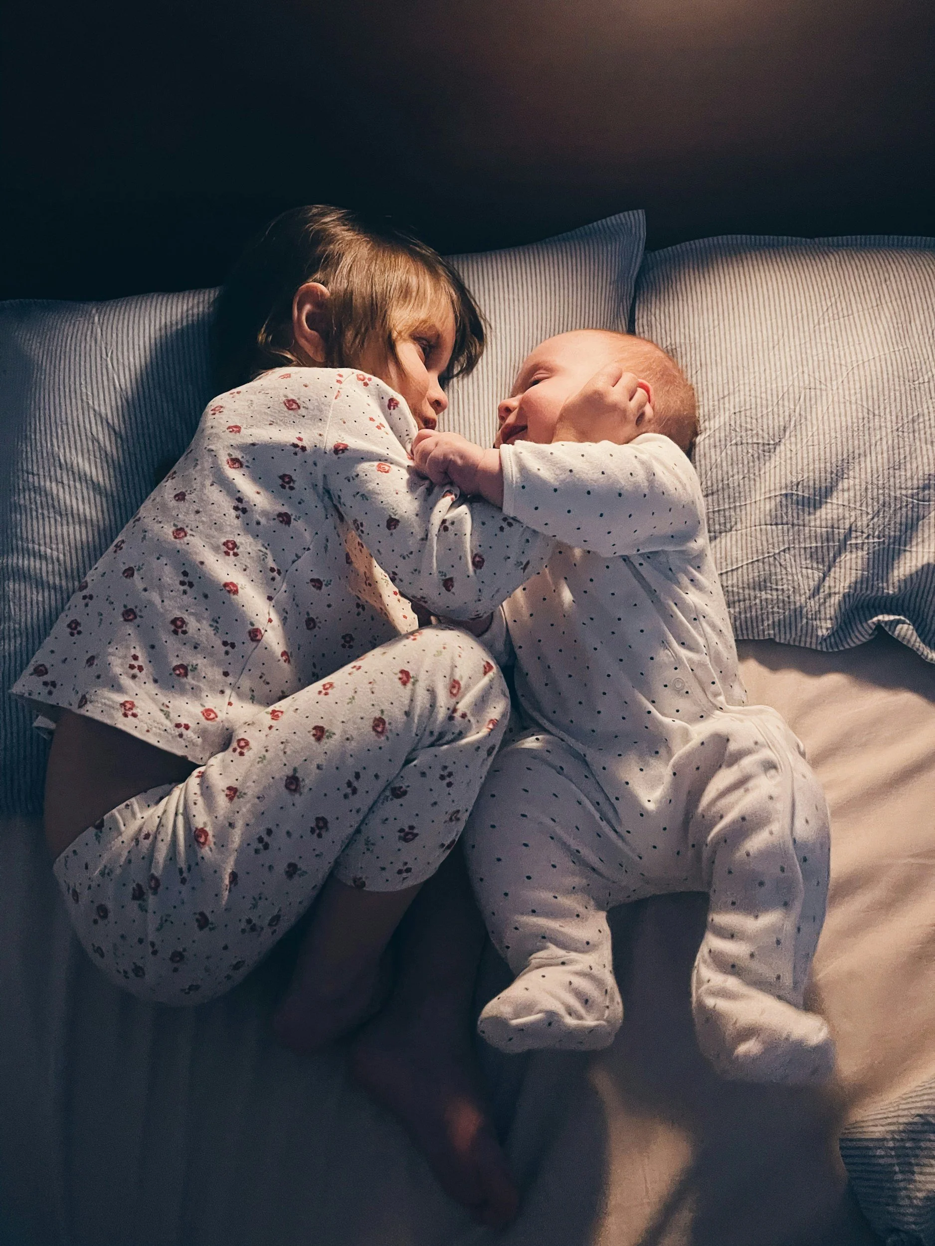 A young girl and a baby lying close together on a bed, facing each other and sharing a moment of affection.