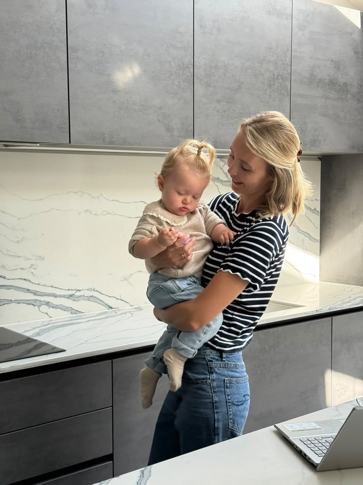 Beth is holding a young girl in a modern kitchen with gray cabinets and a white marble backsplash, engaging with each other.