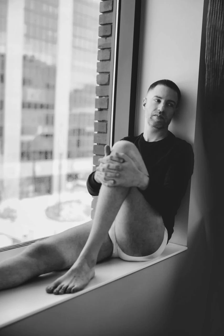 A man sitting on a windowsill, looking out the window, in black and white. He is wearing a dark shirt and white underwear with one knee drawn up and hands holding it.