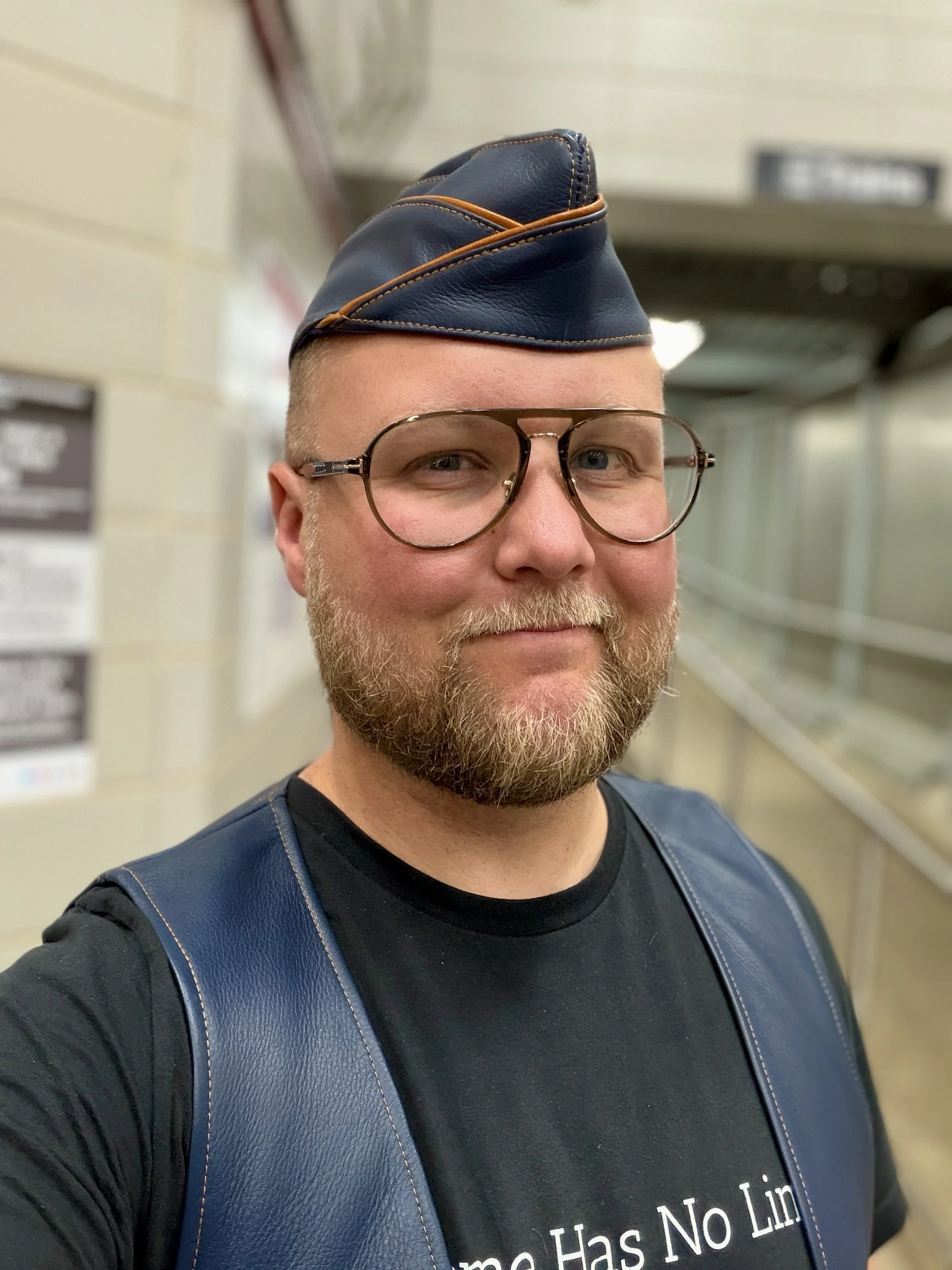 A man wearing glasses and a dark t-shirt with white text, and a navy blue hat with brown piping, standing inside a building with beige walls and shelves in the background.