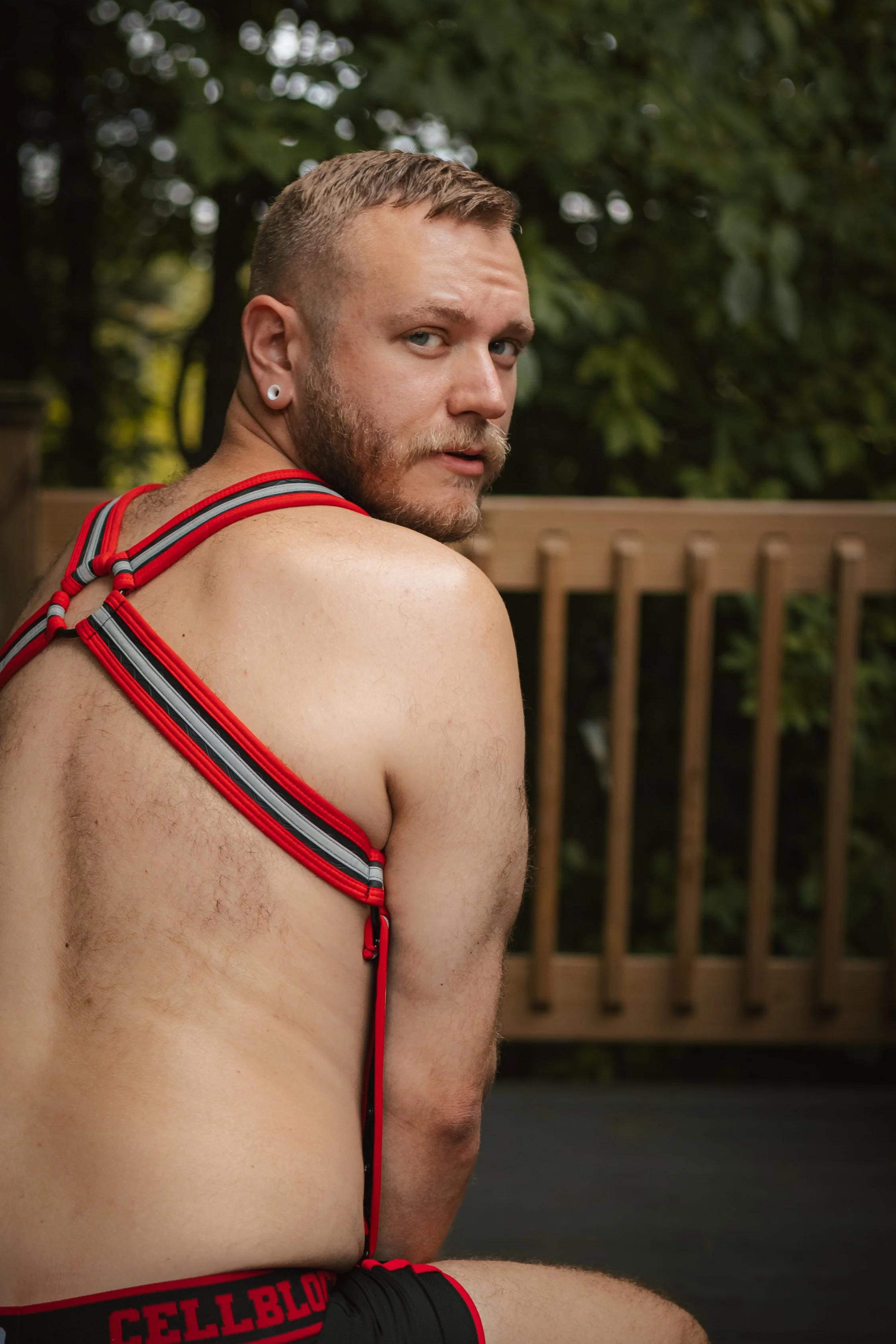 A man with a beard, short hair, and earrings, wearing a red and black harness and black shorts with red trim, sitting outdoors near a wooden fence and green trees.