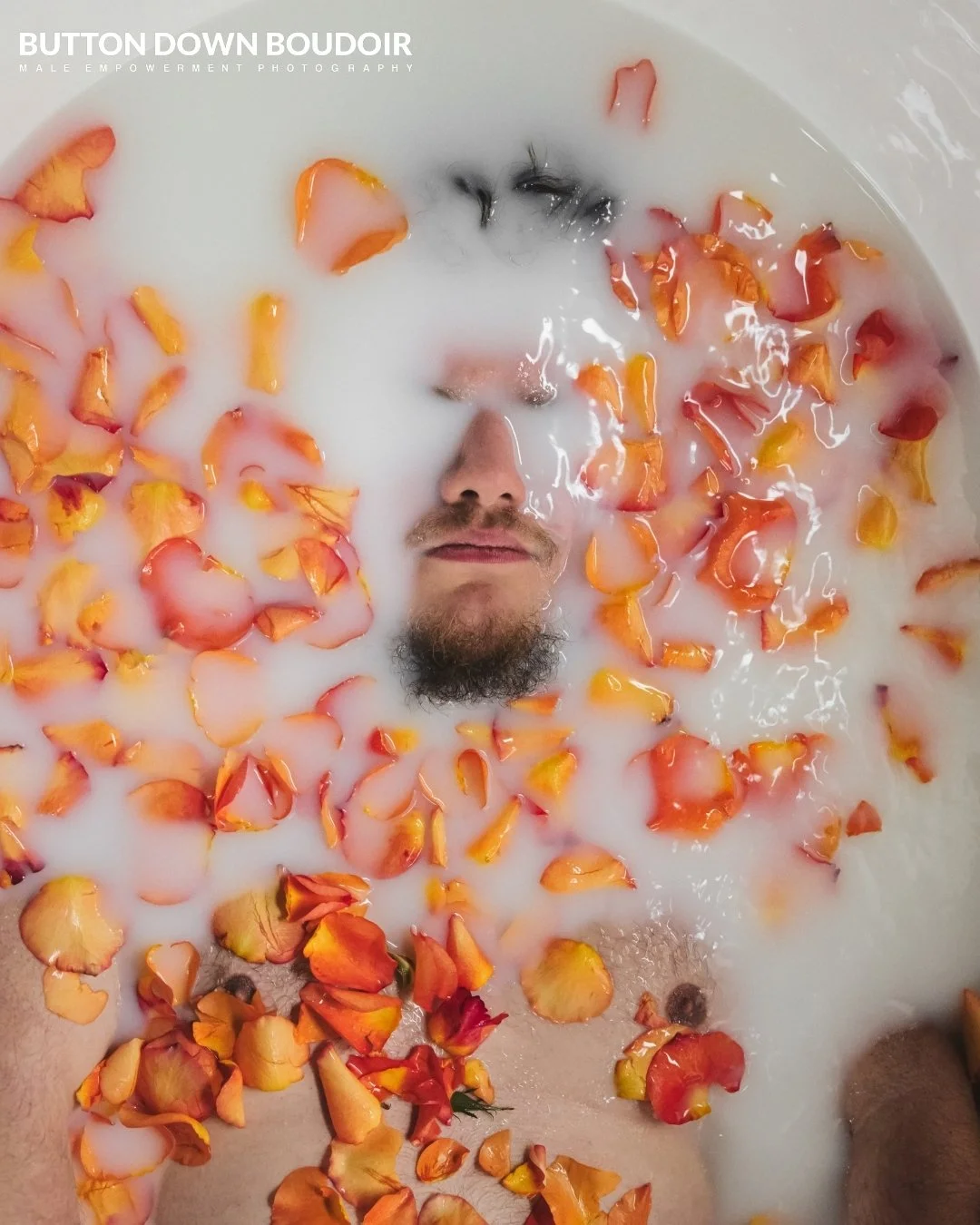 A person with a beard and mustache lying in a bath filled with milk and orange and red rose petals.