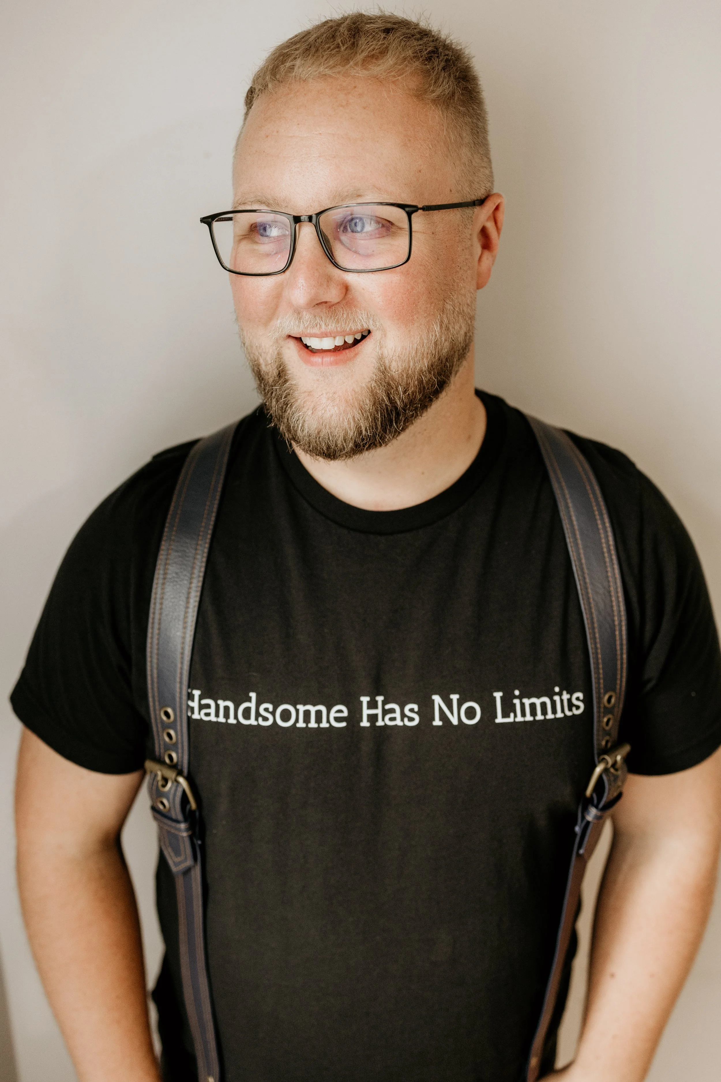 A man with glasses and a beard, wearing a black T-shirt that says "Handsome Has No Limits" and leather suspenders, smiling.