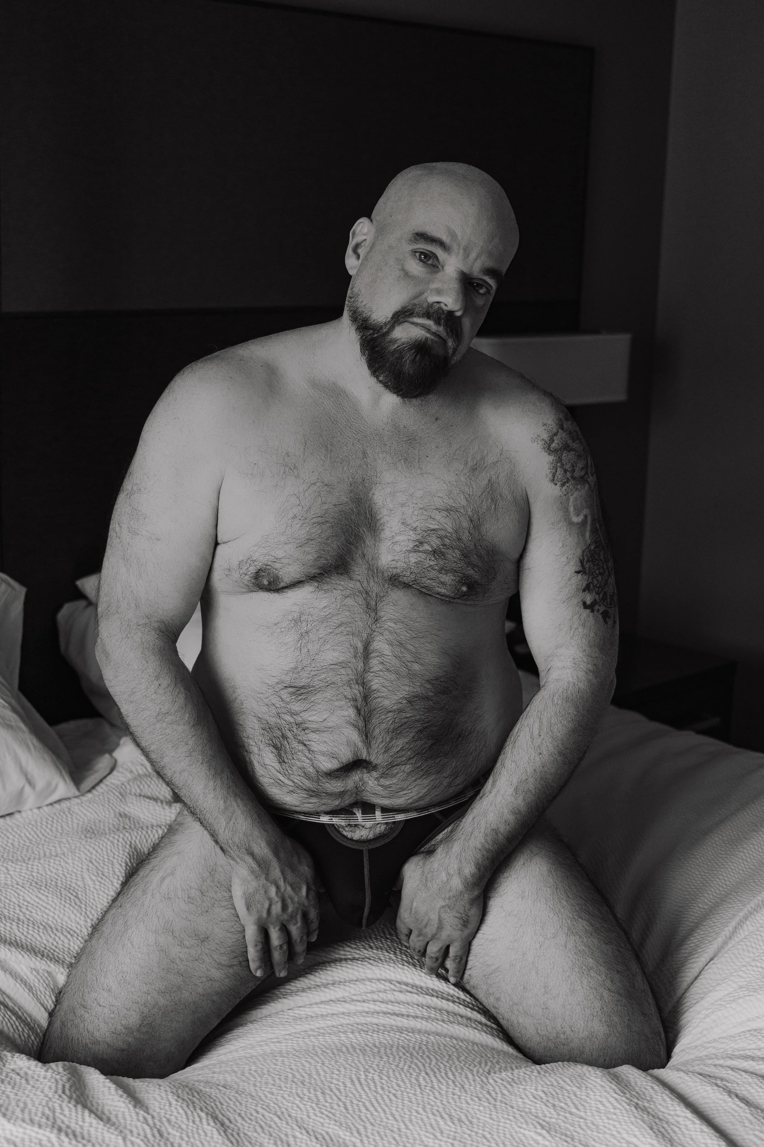 A shirtless man with tattoos on his left arm and a beard, sitting on a bed in a dimly lit room, looking at the camera.