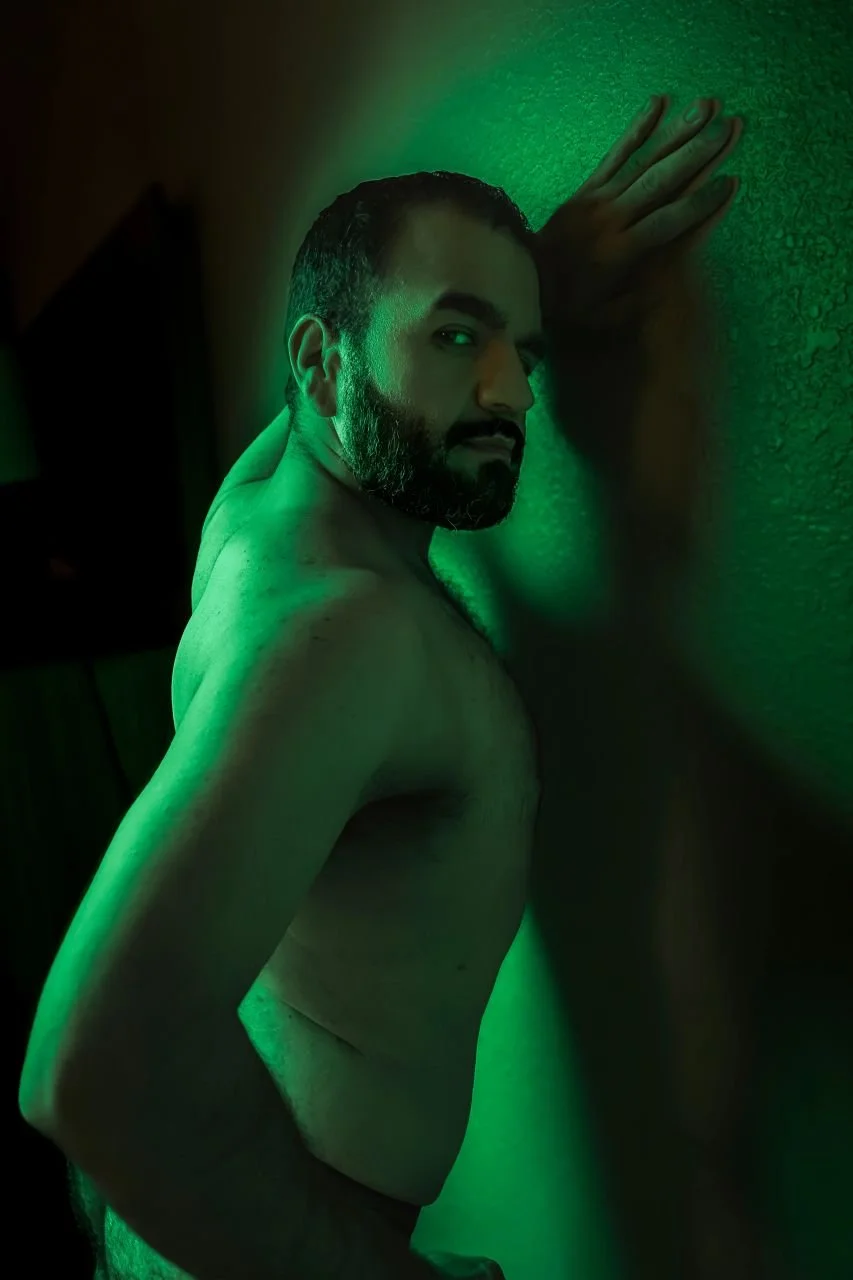 A shirtless man with a beard and short dark hair leaning against a textured green wall, with green lighting creating a moody atmosphere.