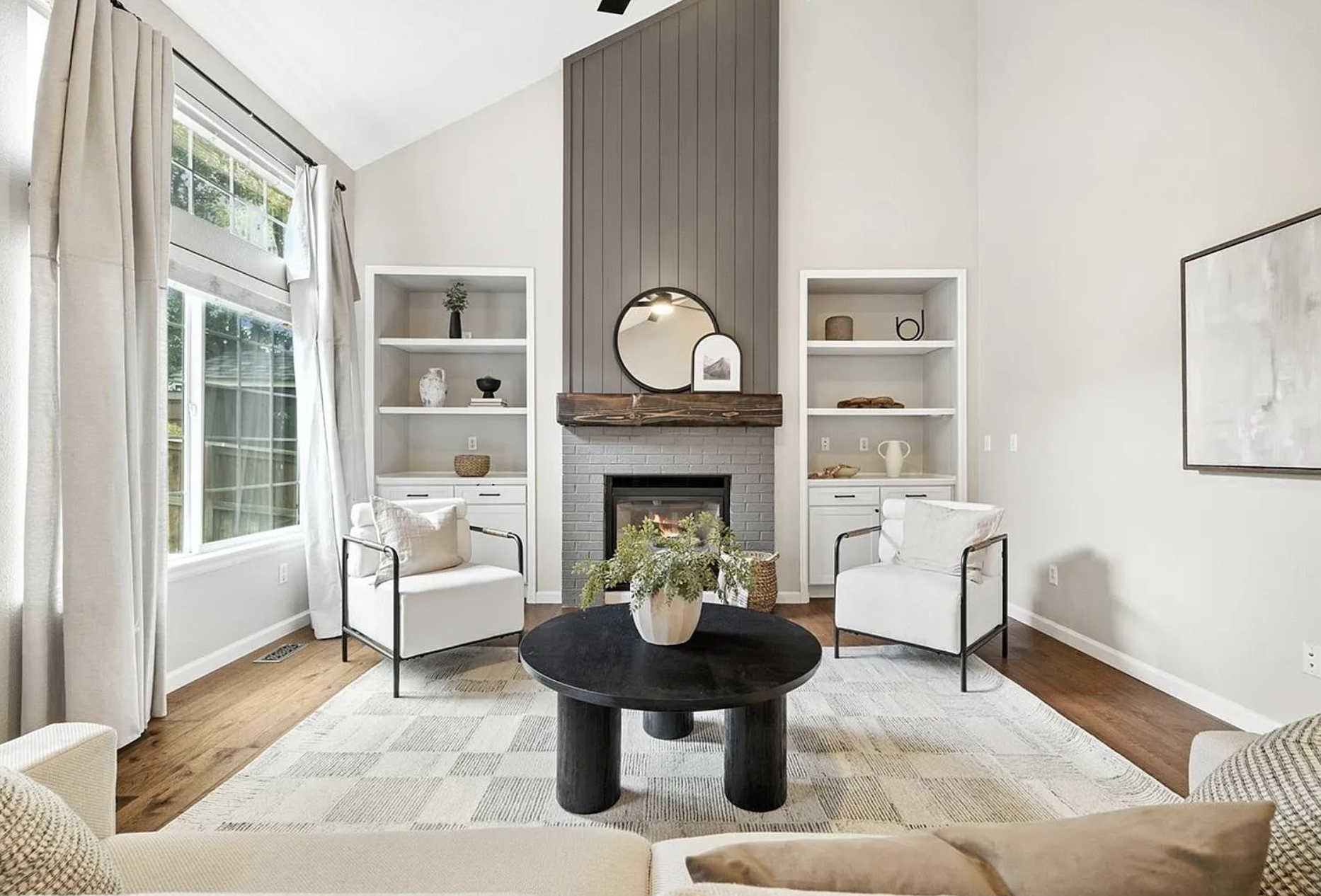 Living room with white walls, hardwood floors, large window with beige curtains, and a fireplace with a dark wood mantel, framed by two built-in white shelves. Two white armchairs with black frames face a black round coffee table with a plant in a white pot. Artwork hangs on the wall to the right.