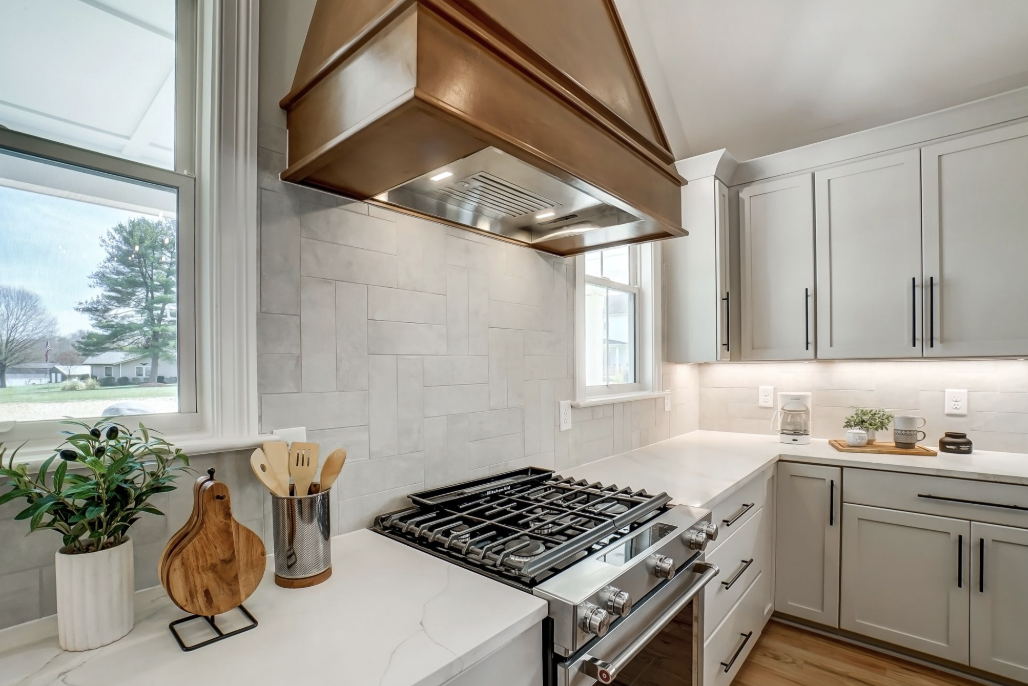 Modern kitchen with white cabinets, a gas stove, a wood-range hood, and a window with a view of trees and houses outside.